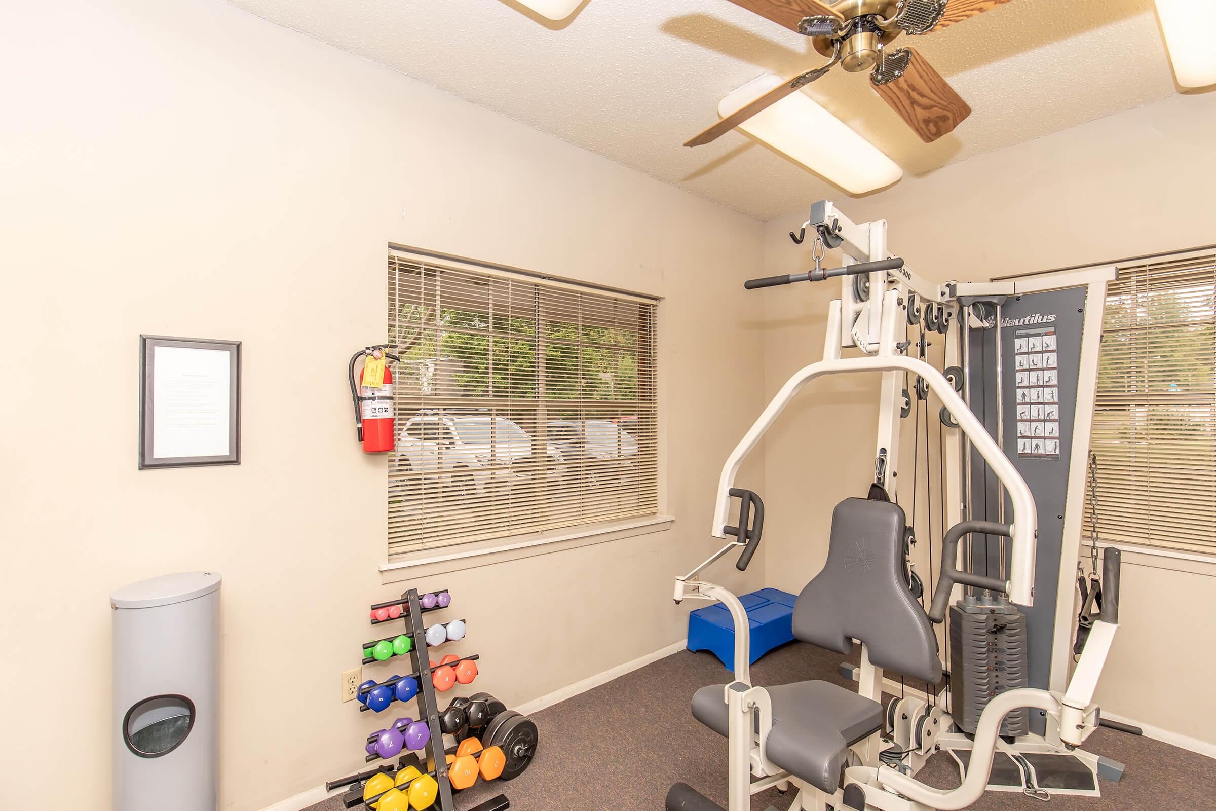 A small gym space featuring a multi-station exercise machine, dumbbell rack with colorful weights, a blue exercise bench, and a window with blinds. There is also a fire extinguisher mounted on the wall and a trash can in the corner, along with a framed document on the wall.