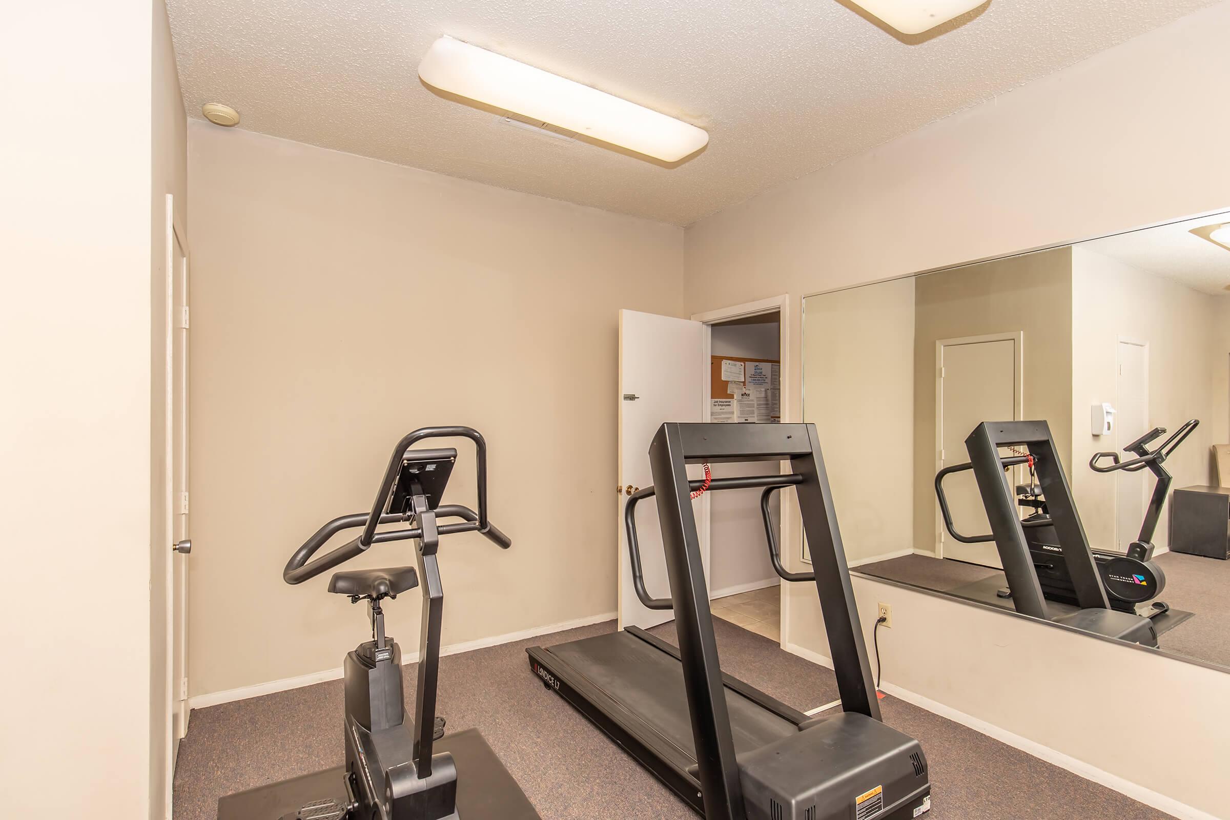 A small, simple gym space featuring a treadmill and a stationary bike. The room has beige walls and a large mirror that reflects the equipment. There is a door on the right leading to another area, and basic overhead lighting provides illumination. The floor is carpeted, contributing to a cozy atmosphere.