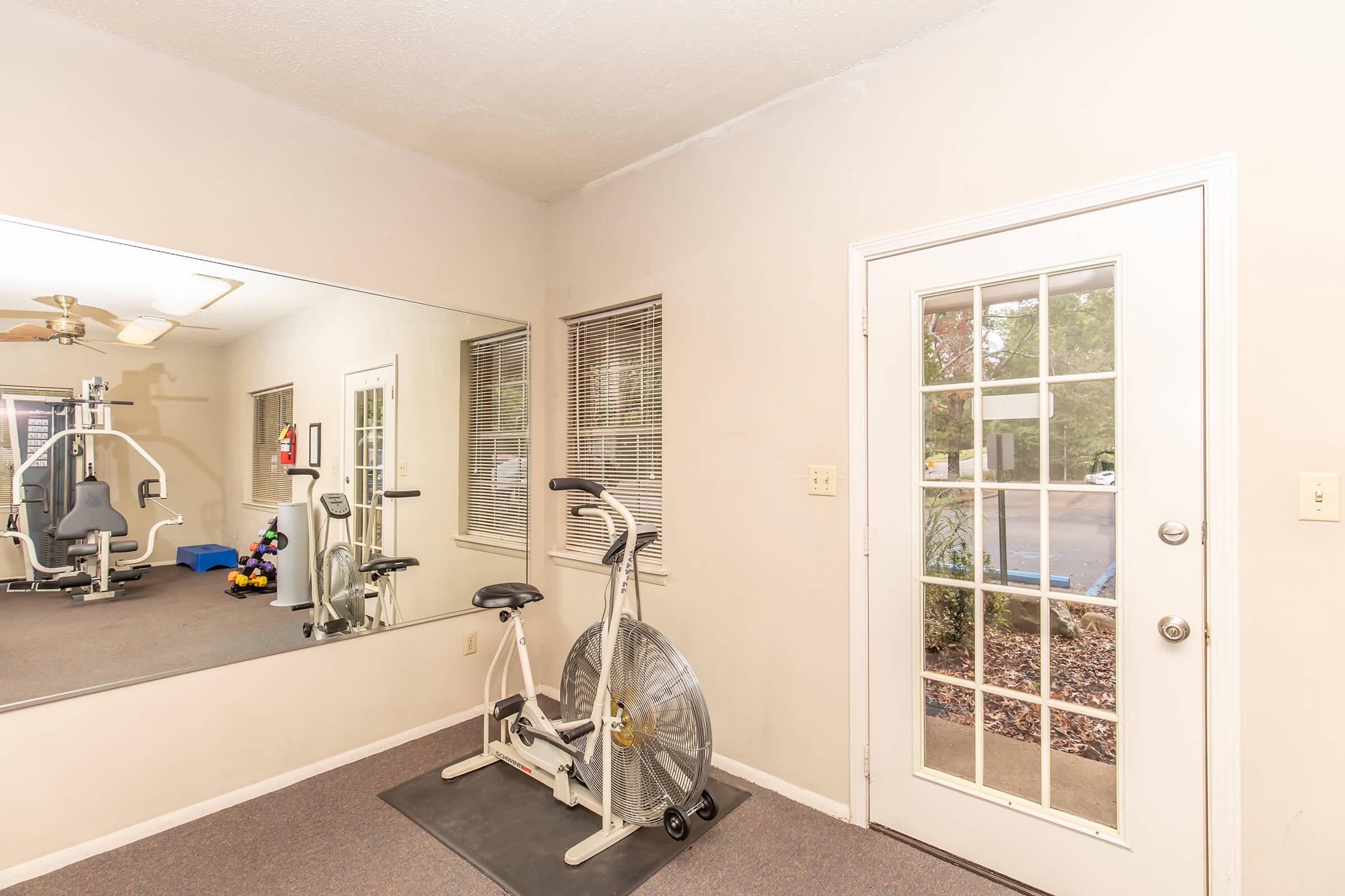 Interior of a gym space featuring a stationary exercise bike in front of a large mirror. A door leads outside, with natural light coming through. On the floor are exercise weights and fitness equipment in the background. The walls are light-colored, creating an open atmosphere.