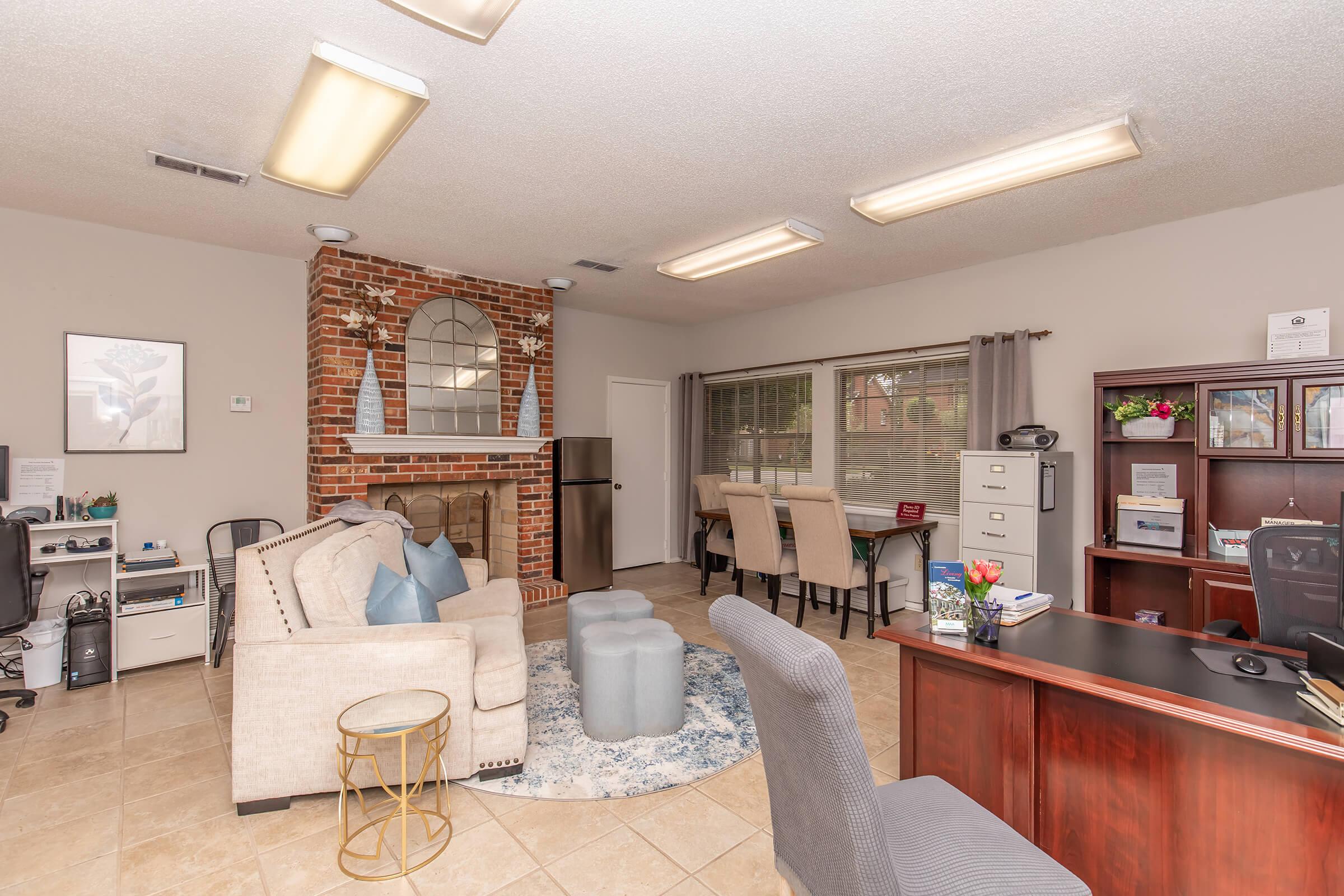 A cozy office space featuring a beige sofa with blue and white accents, a circular area rug, a decorative brick wall with a mirror, and a desk with a computer. There are plants, a stylish chair, and a dining table with chairs in the background, all creating a welcoming atmosphere.