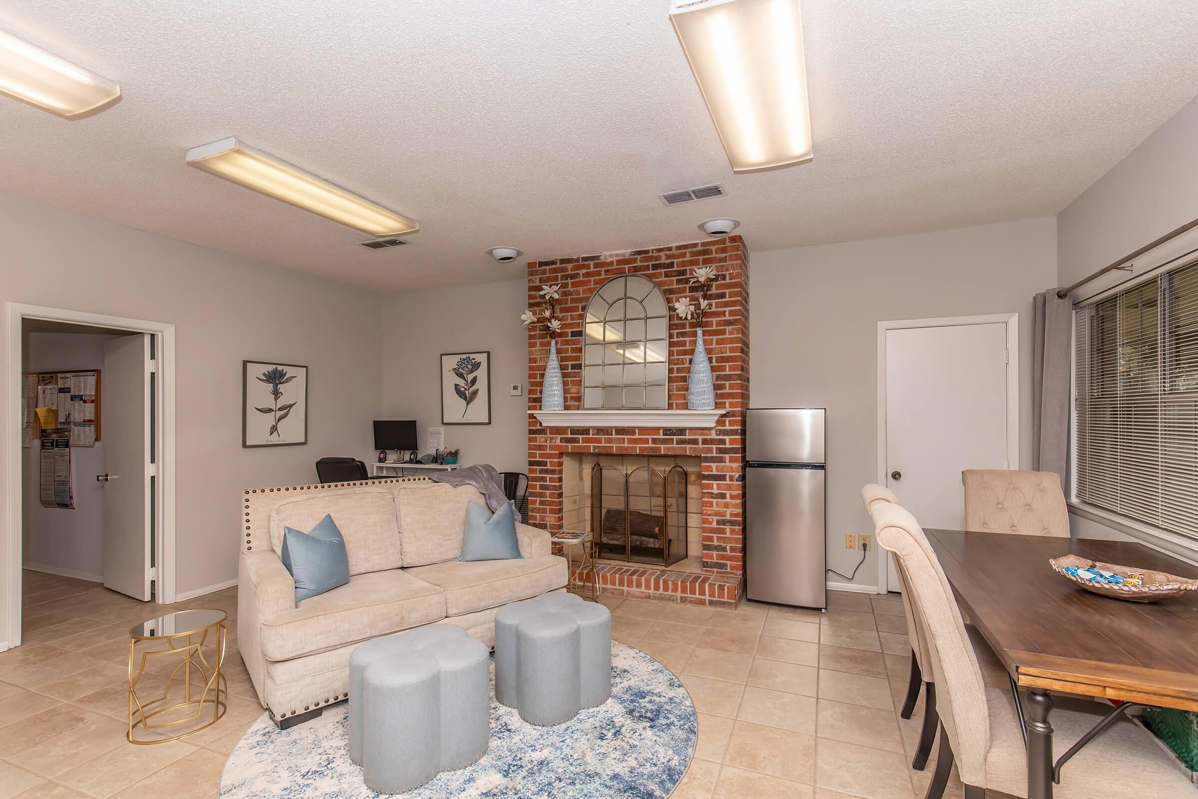 Cozy living space featuring a beige sofa with blue cushions, a decorative fireplace with a brick design, and a round area rug. There is a dining table with chairs, a refrigerator, and a workspace with a computer setup. Bright overhead lights illuminate the room, creating a welcoming atmosphere.