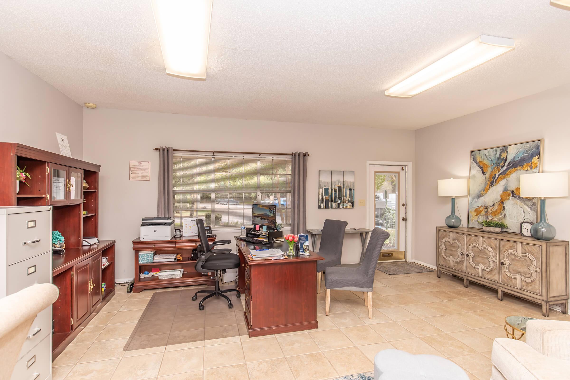 A tidy office space featuring a desk with a computer, a bookshelf, and a filing cabinet. Two gray upholstered chairs are positioned around a small table. There's a large window letting in natural light, with curtains providing a soft touch. Decorative artwork and lamps enhance the welcoming atmosphere.