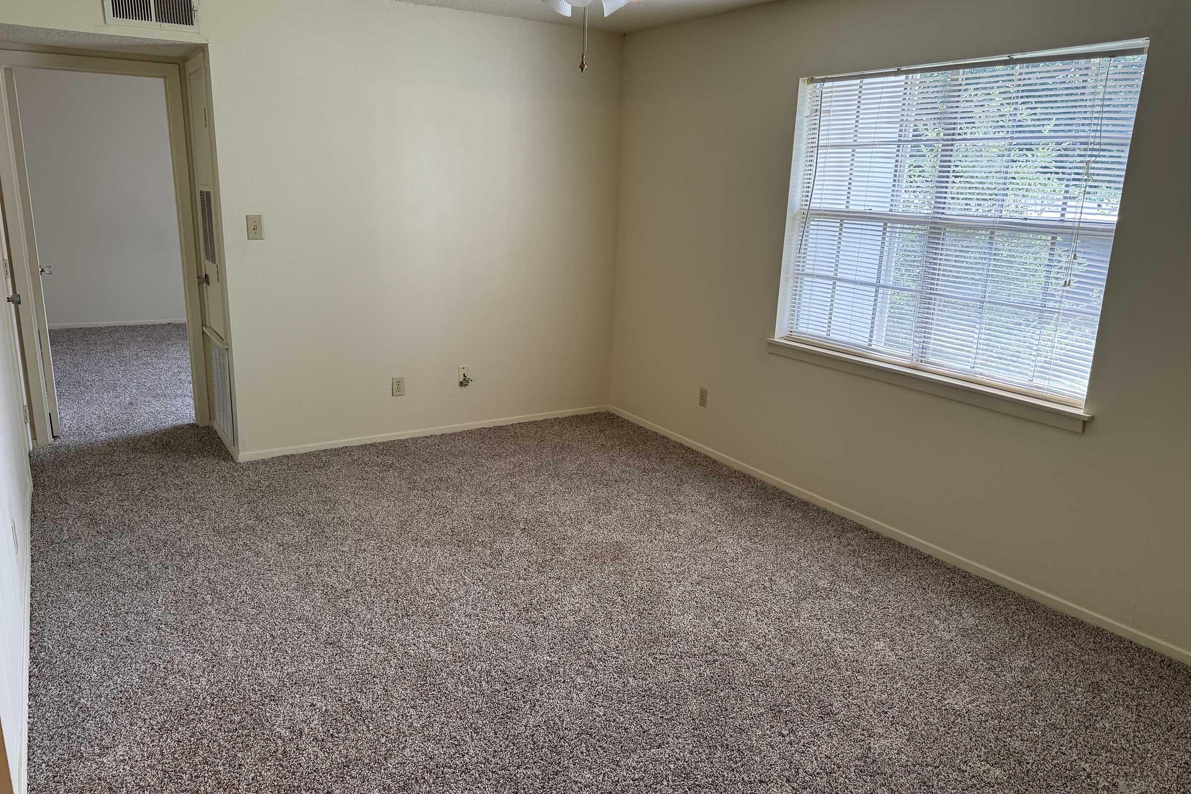 A vacant room with beige walls and light-colored carpet. The room features a window with blinds allowing natural light, and a ceiling fan. There are two doorways: one leading to another room and the other likely to a hallway.