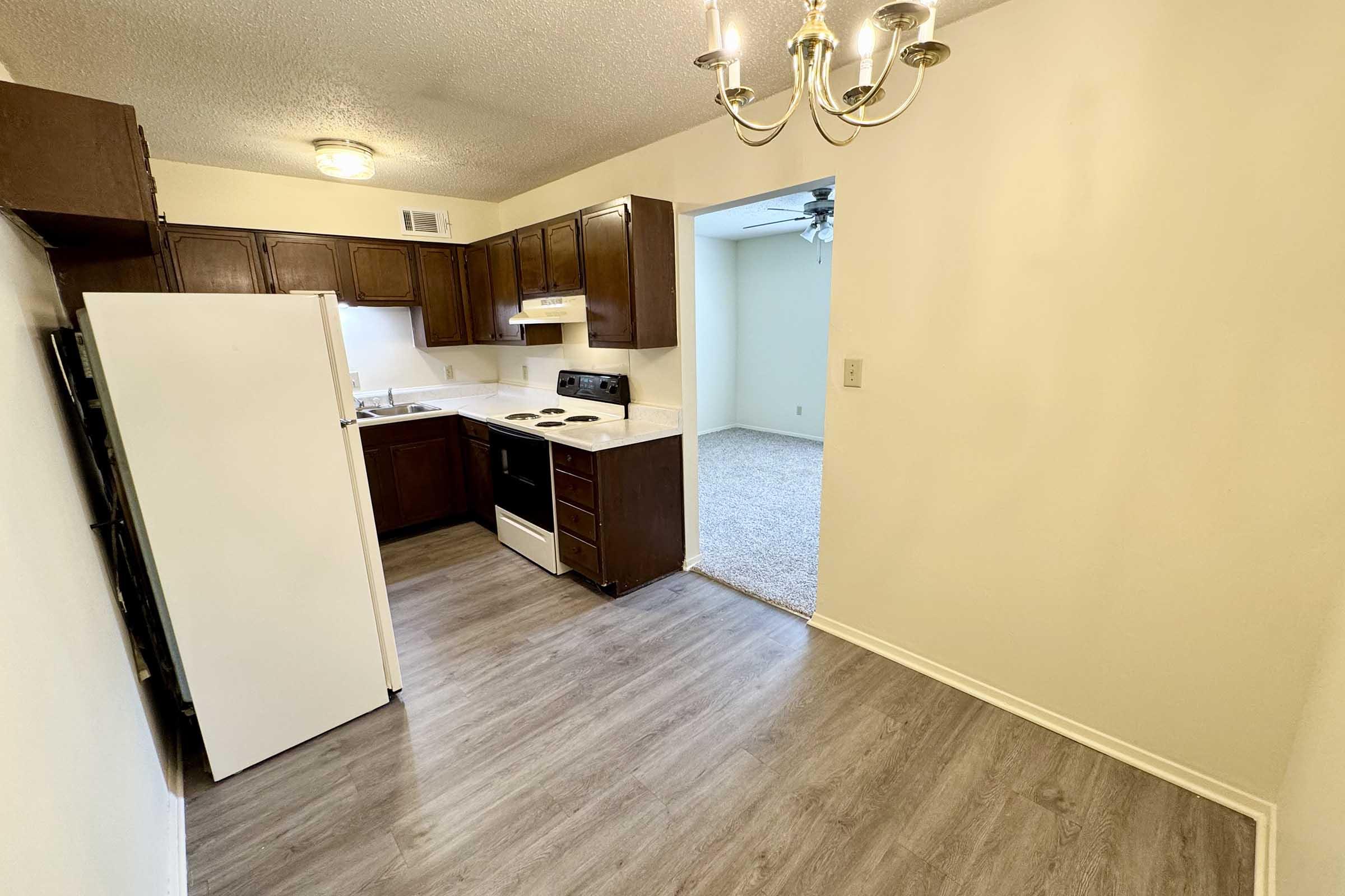 A small kitchen featuring dark wooden cabinets, a white refrigerator, and an oven with a stovetop. The floor is light-colored laminate, and there's a chandelier above. An open space leads to a carpeted living area in the background.