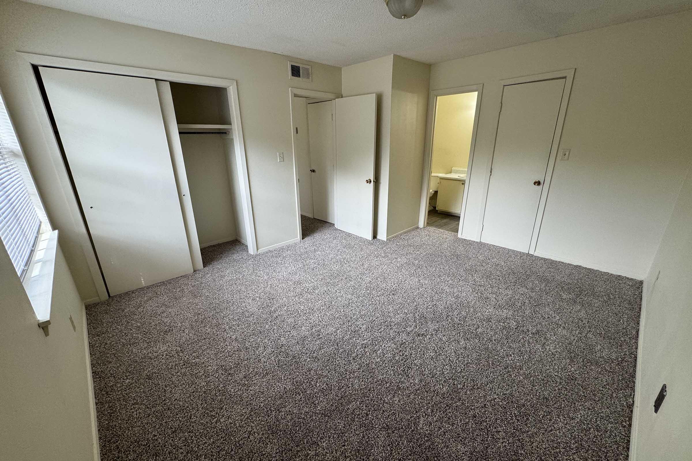 A spacious, empty room with light-colored walls and beige carpeting. Two closed closet doors line one wall, and a doorway leads to an adjacent room. A single light fixture hangs from the ceiling, providing overhead illumination. Windows allow natural light to enter, highlighting the room's clean and unoccupied appearance.