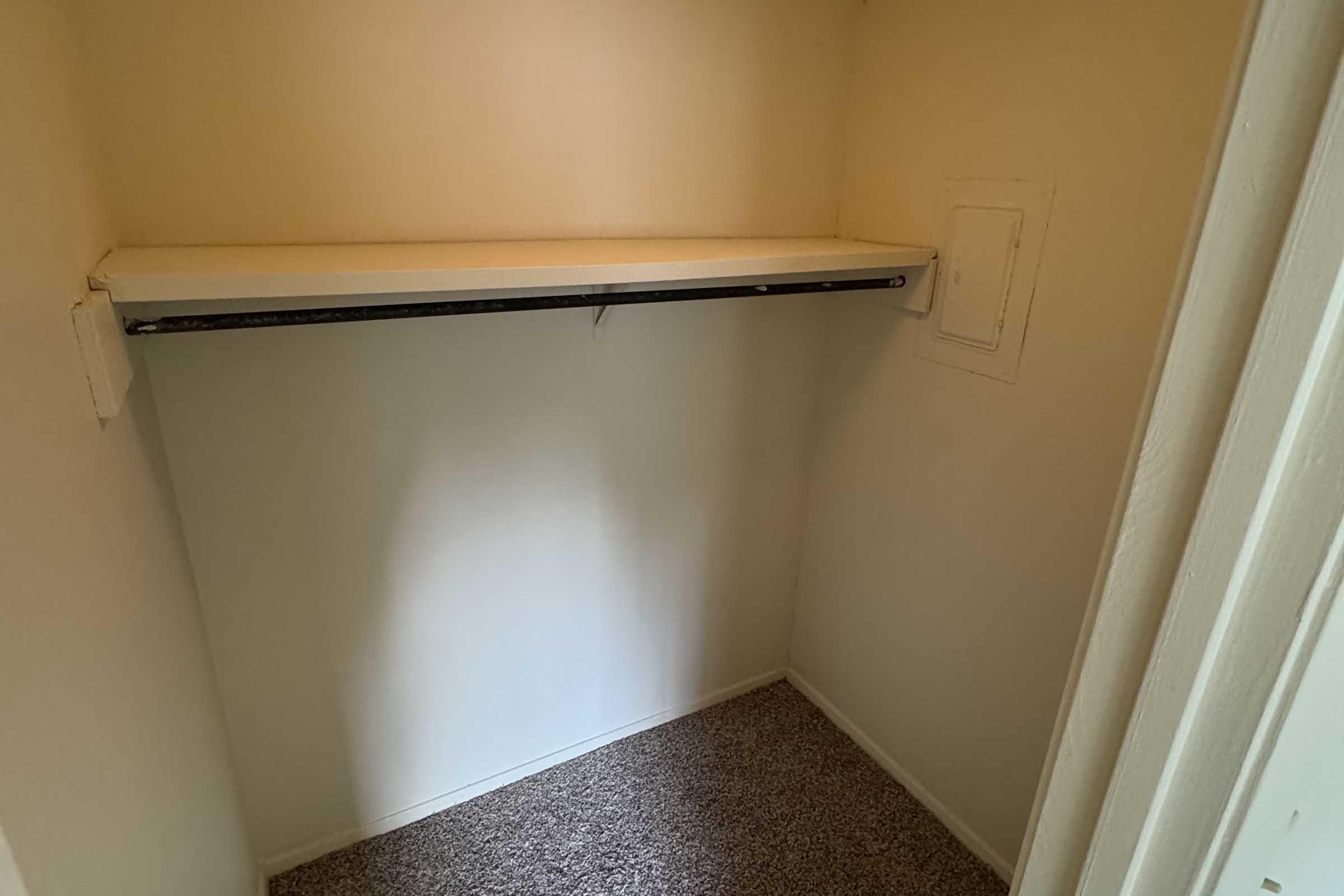 Empty closet with a white shelf along the top and a clothing rod beneath it. The walls are painted a light color, and the floor is covered with a tan carpet. There is a small, closed opening in one wall, possibly for access to utilities.