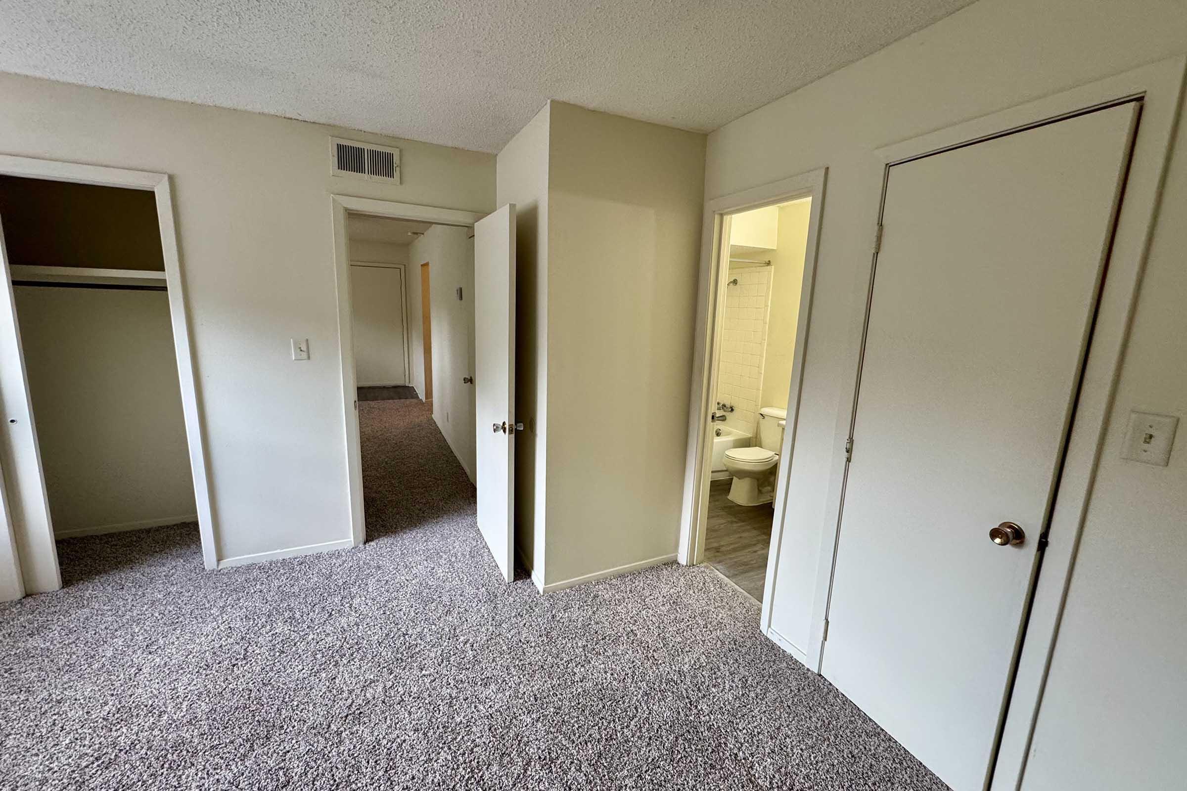 A view of a small, carpeted room featuring multiple doors leading to a closet and a bathroom. The walls are painted in neutral colors, and the space appears well-lit with a doorway visible in the background.