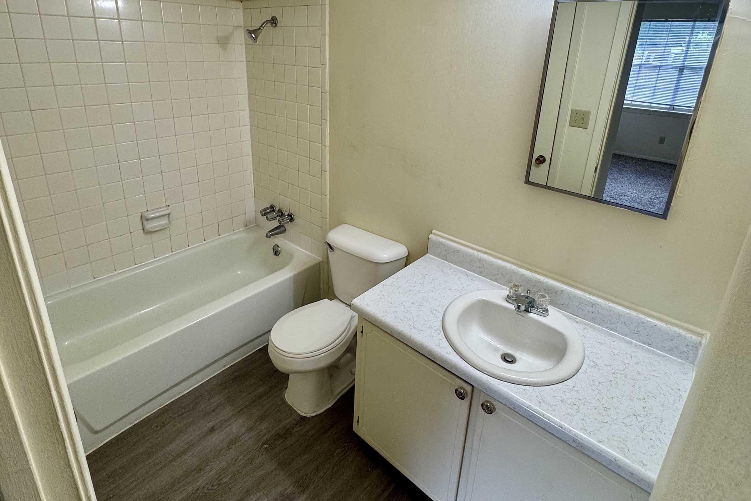 A small bathroom featuring a bathtub with a shower, a toilet, and a sink with a countertop. The walls have light-colored tiles, and there is a mirror above the sink. A window provides natural light, illuminating the space with neutral-colored walls and flooring.