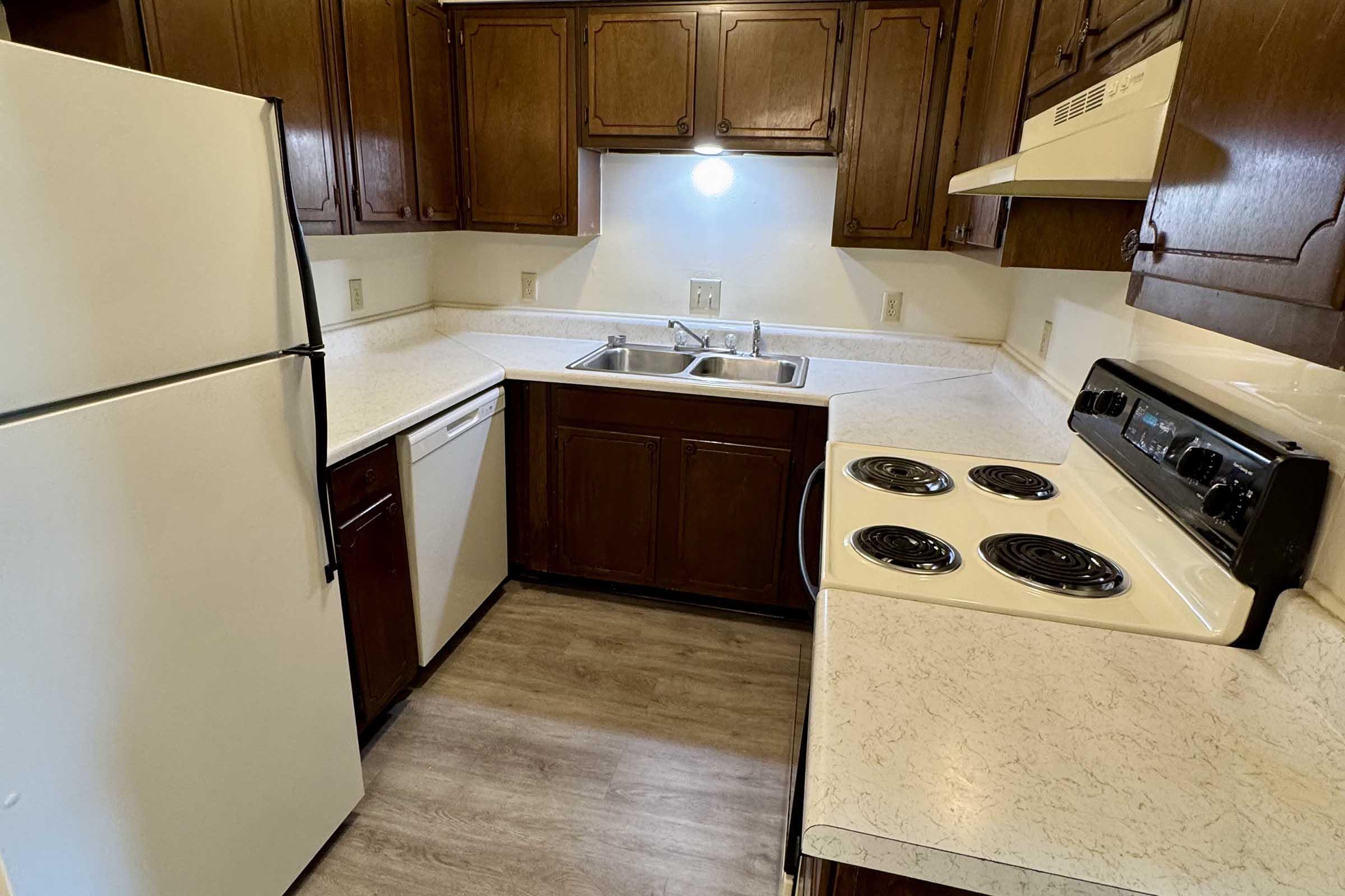 A compact kitchen featuring wooden cabinetry, a white refrigerator, a dishwasher, a double sink, and a black stove with four burners. The countertops are light-colored, and the floor is a neutral wood-like laminate.