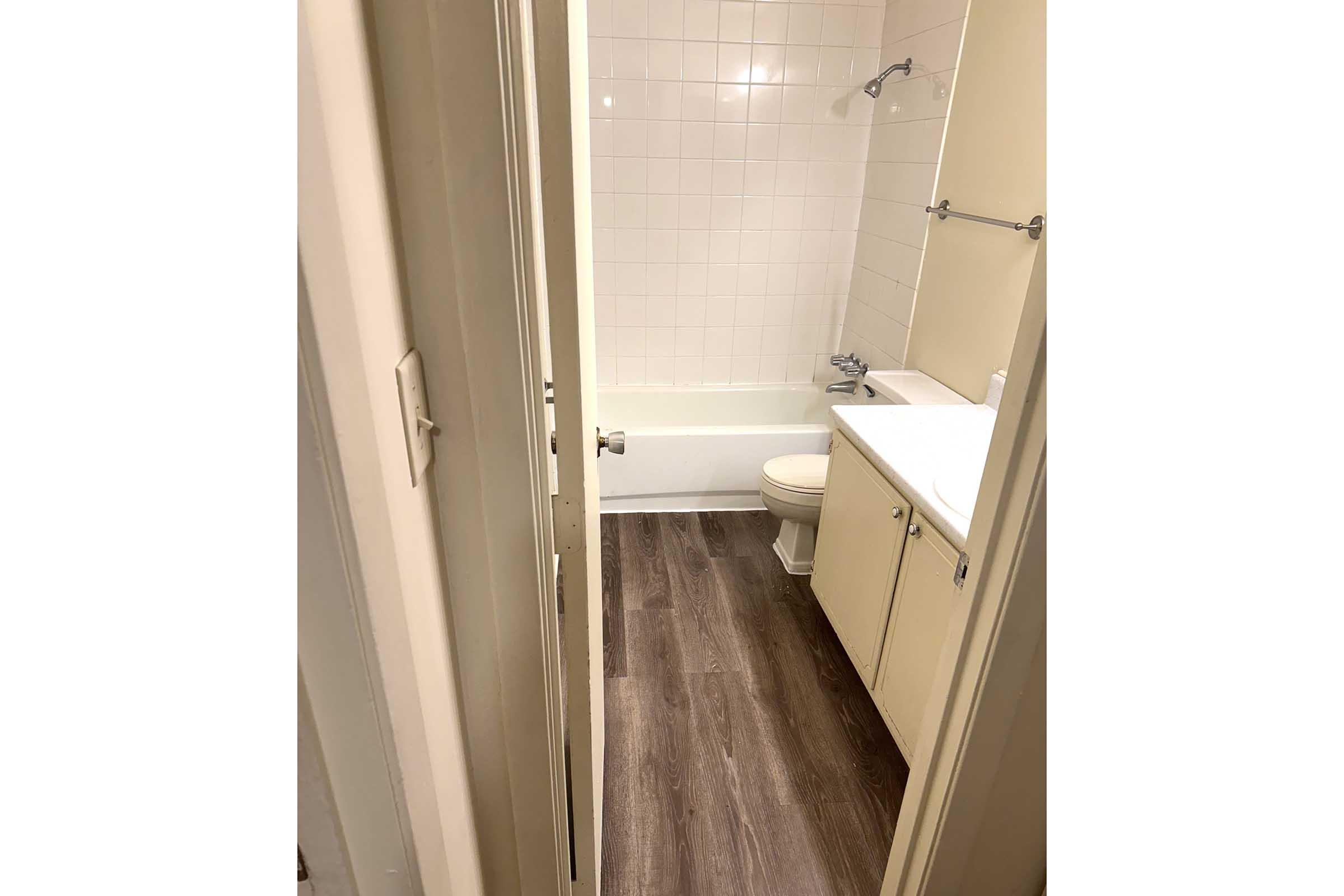 A view of a small bathroom, featuring a bathtub, toilet, and sink. The walls are tiled, and the floor has wooden laminate. There is a doorway leading to the bathroom, showing a glimpse of the interior design and layout.