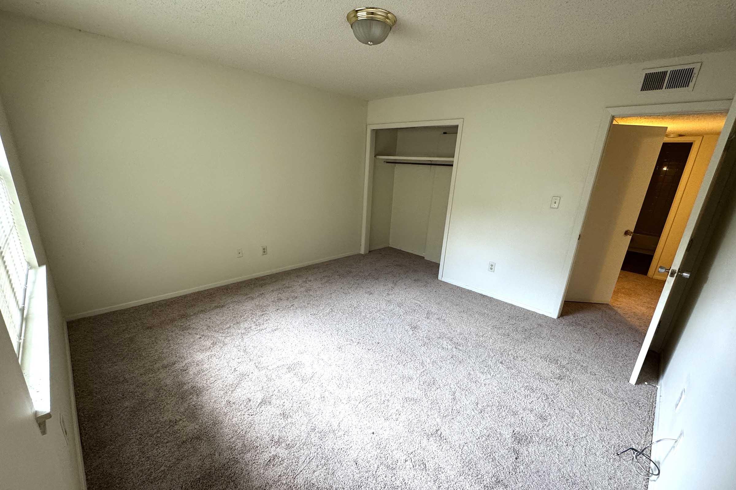 A sparsely furnished, empty bedroom with light beige walls and carpet. There is a window allowing natural light, a closet with sliding doors, and an open door leading to another room or hallway. The overall atmosphere is bright and clean, suggesting the space is ready for new occupants.