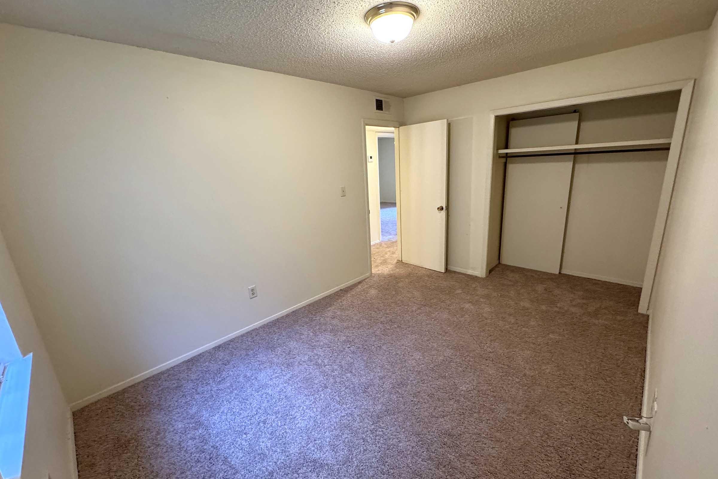 Empty room with light beige walls and carpeting. A closet is visible on the right side, and there is an open doorway leading to another room. The ceiling features a round light fixture, and the natural light enters from a window on the left. The overall space appears clean and well-maintained.