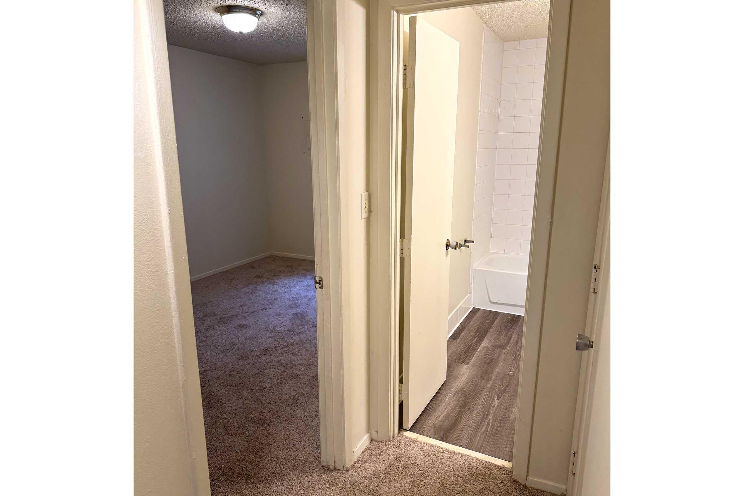 A view of an interior hallway leading to two doors. One door opens to a room with carpeted flooring, while the other leads to a bathroom featuring a tub and tiled walls. The walls are painted in neutral colors, and there is a ceiling light fixture visible.