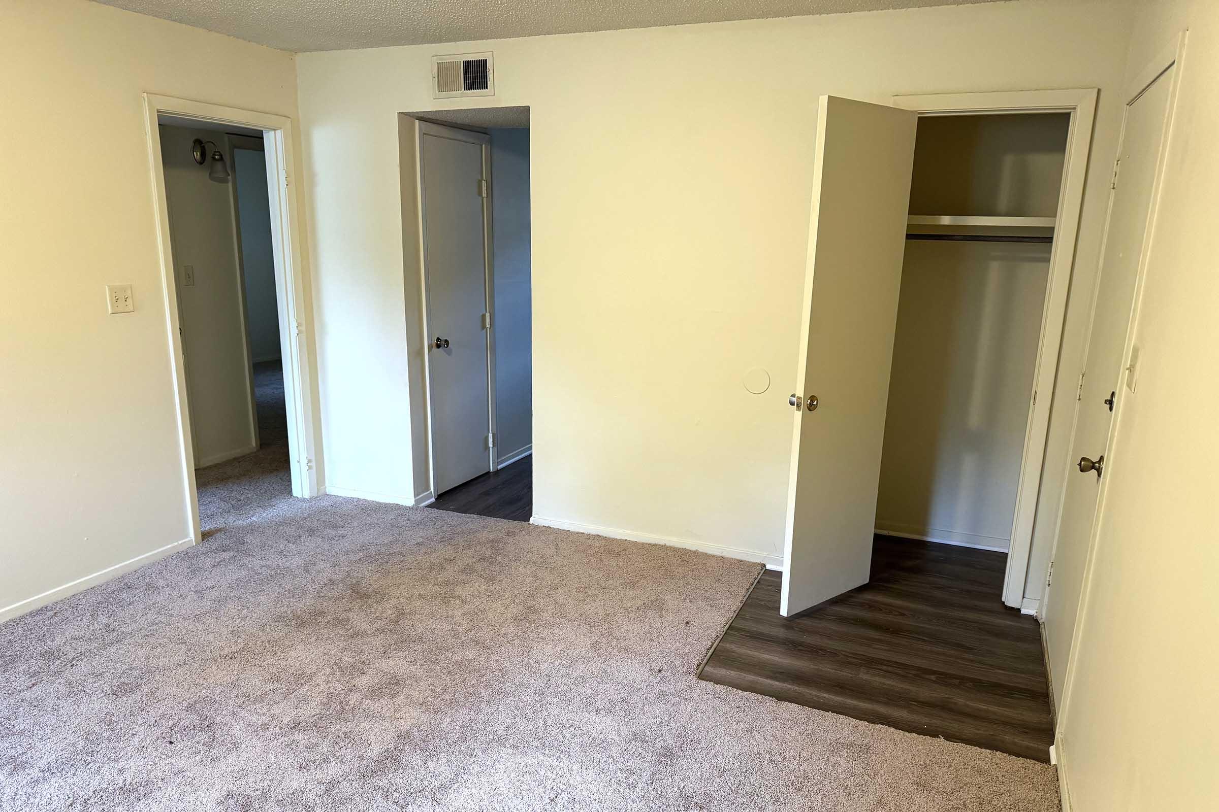 A view of a softly lit room featuring light-colored walls and carpeted flooring. There are two doorways leading to adjacent rooms, with one partially open revealing a closet. The floor transitions from carpet to laminate near the closet entrance.