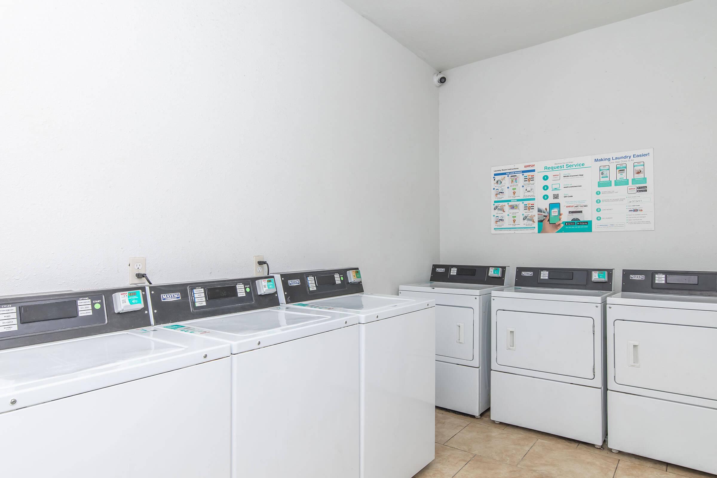 A clean, well-lit laundry room featuring multiple white washing machines and dryers. The machines are lined up against a light-colored wall, and there is a poster on the wall displaying laundry instructions and tips. The floor is tiled, and the overall atmosphere is organized and functional.