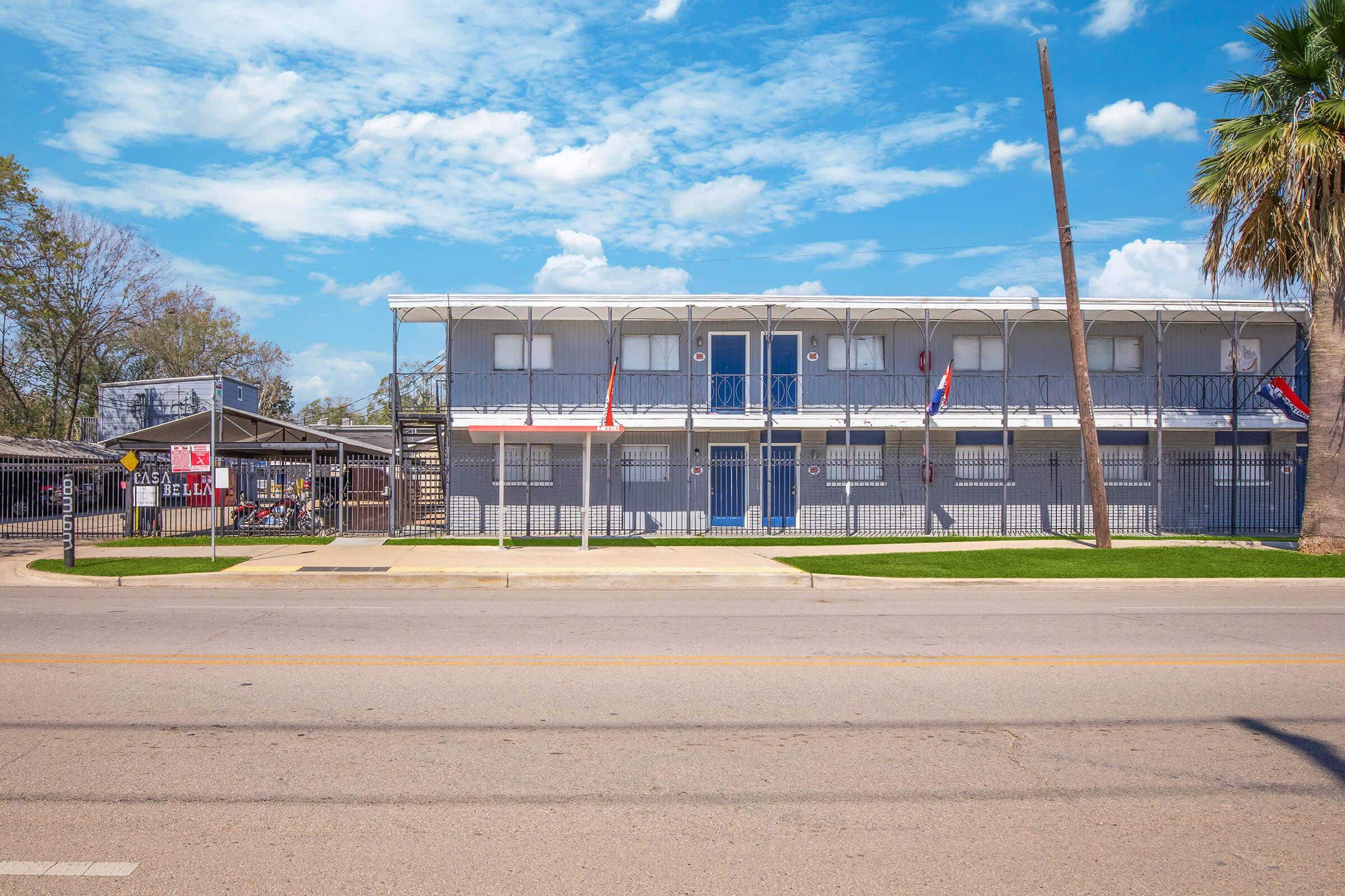A two-story building with a white exterior, featuring blue balconies and several flags hanging from it. In the foreground, there is a paved street with a grassy area. The sky is clear with a few clouds, and a palm tree is visible on the right side.