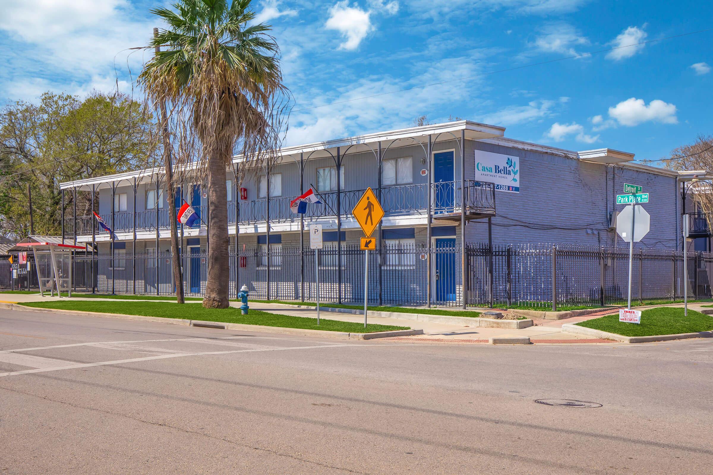 A low-rise building with multiple balconies, featuring a blue exterior and palm trees in front. There are flags displayed and a street sign indicating a pedestrian crossing. The scene is set against a clear blue sky with scattered clouds.
