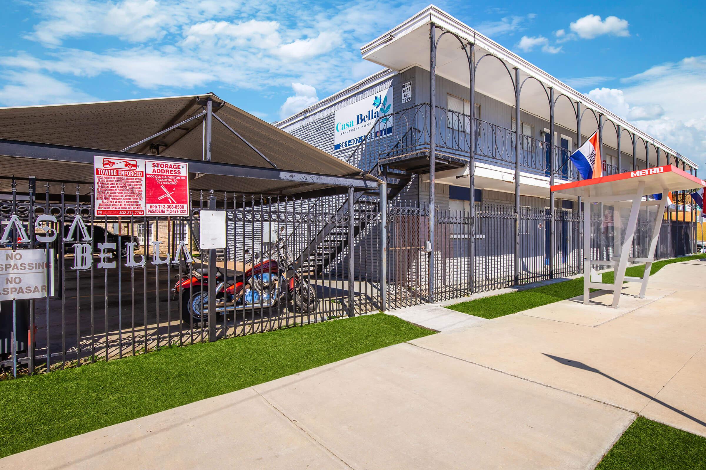 Exterior view of a multi-story building with a sign reading "Casa Bella." The entrance features a covered area with fences and signage, including warnings about security and trespassing. A motorcycle is parked nearby, and there is a bus stop shelter on the sidewalk. The scene is set under a clear blue sky.