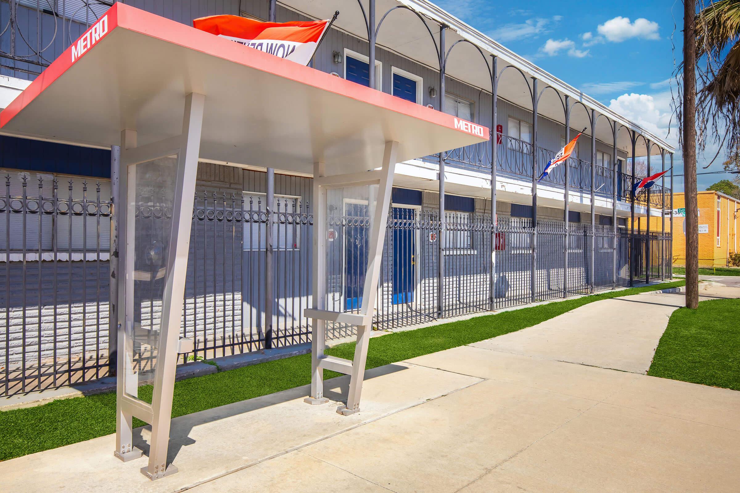 A bus stop shelter with a red canopy sits on a sidewalk next to a building with a blue facade and multiple balconies. The area is well-lit, featuring green grass along the sidewalk and several flags visible on the property. Clear skies provide a bright atmosphere.