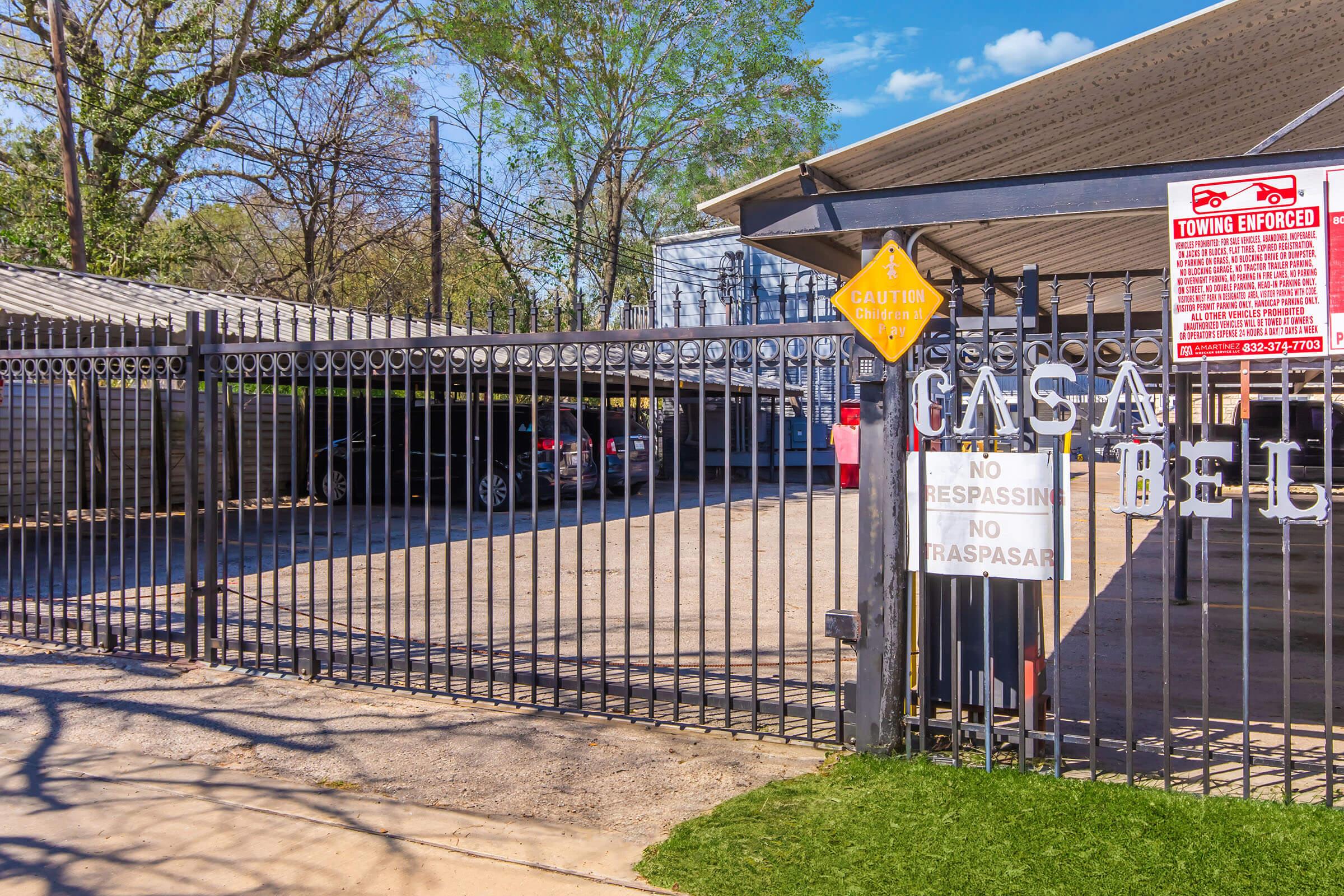 A gated parking area with a chain-link fence. Signs indicate "Caution" and "No Trespassing," along with a towing enforcement notice. The area is surrounded by trees and features a concrete surface. A car is partially visible in the background under the covered parking.