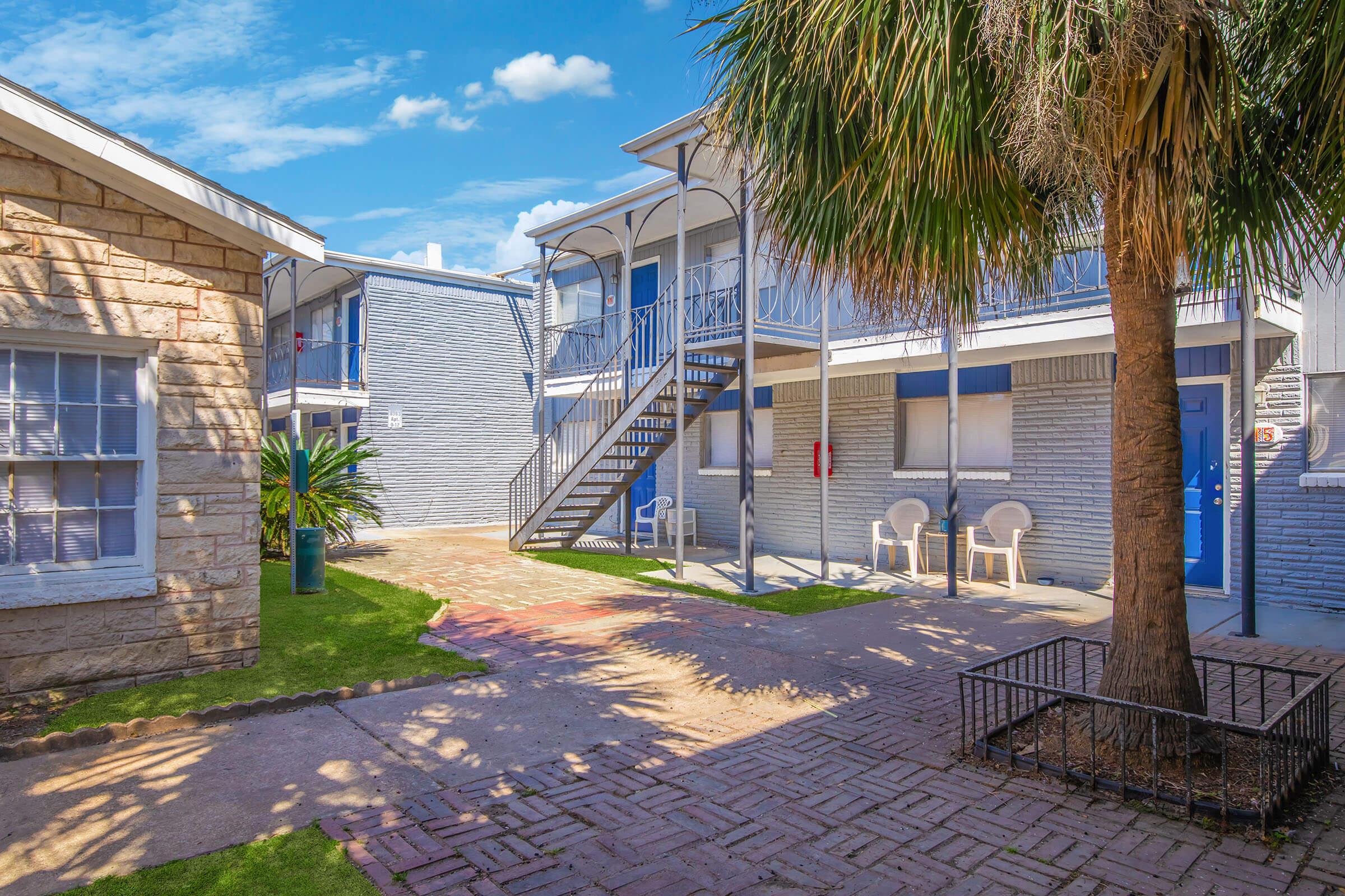 View of a well-lit courtyard featuring two multi-story buildings. One building has a staircase leading to upper apartments, while the other has stone walls. Lush greenery, including a palm tree, surrounds the area, with chairs positioned outside and a blue sky overhead.