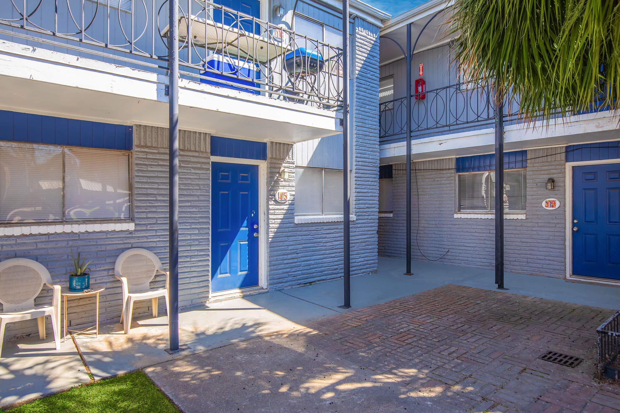 A view of a courtyard between two blue doors of an apartment building. The area features a small seating arrangement with two white chairs and a potted plant, along with a sunlit pathway and textured walls. The setting is clean and well-maintained, surrounded by tropical foliage.