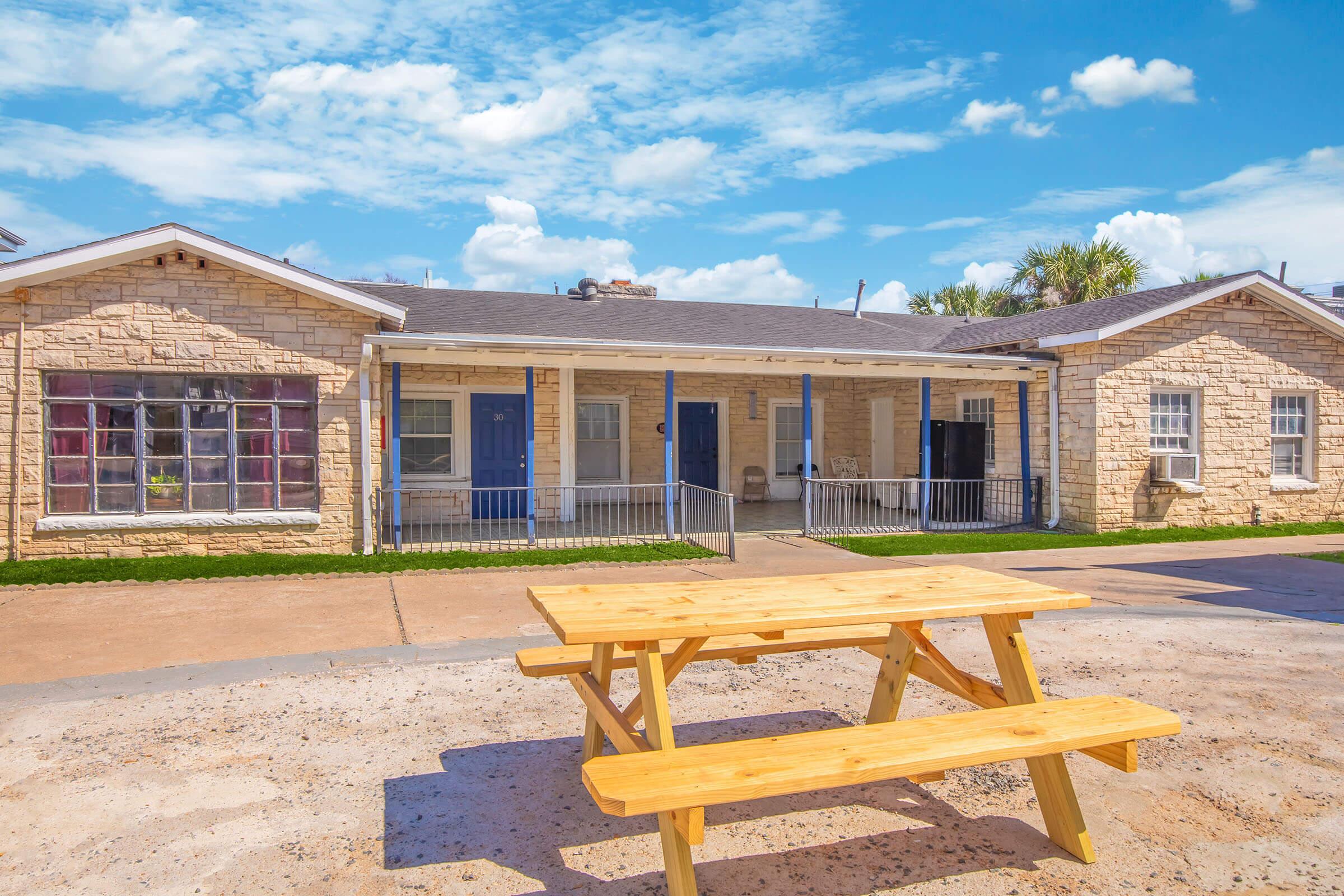 A view of a light-colored stone building with several doors, situated under a bright blue sky with scattered clouds. In the foreground, there is a wooden picnic table on a gravel surface, indicating a casual outdoor setting. The area appears well-maintained and inviting.
