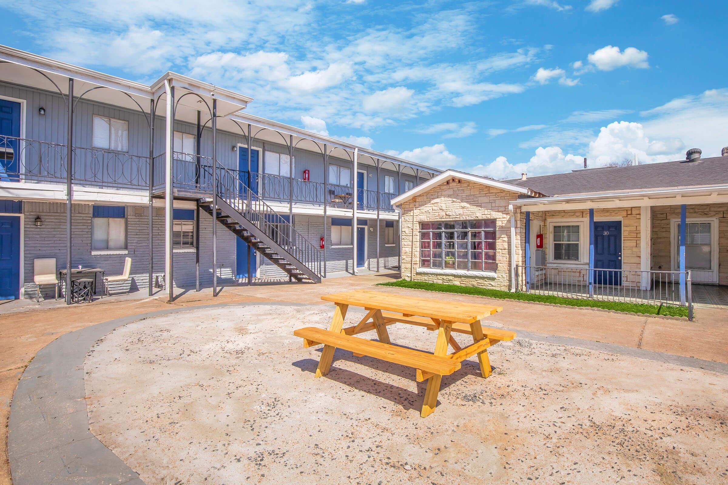 A courtyard view of a motel with two levels of rooms, featuring a wooden picnic table in the center. There are blue doors on the rooms, a staircase leading to the upper level, and a building with large windows. The sky is bright with scattered clouds, and the ground is made of gravel and pavement.