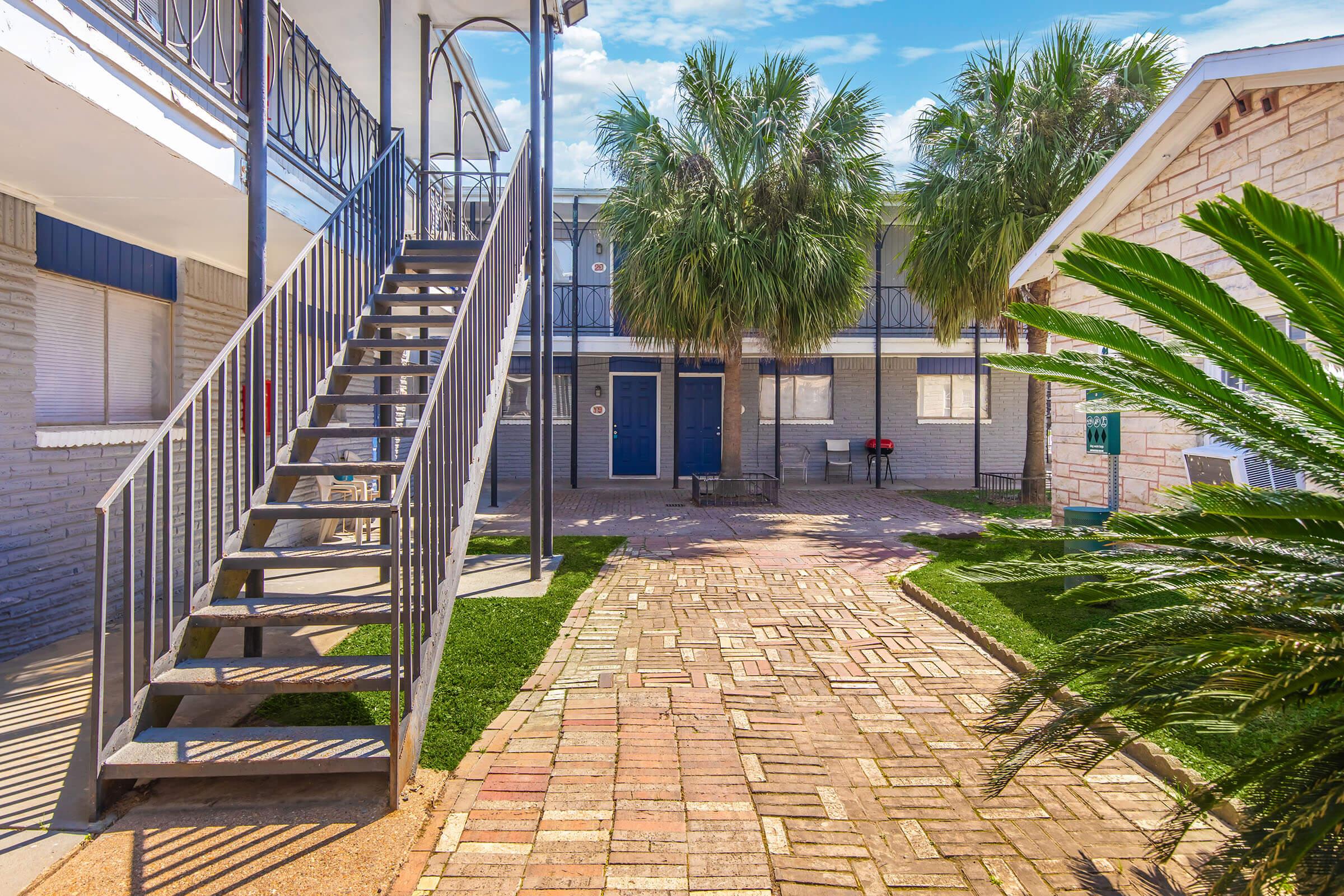 An outdoor view of a multi-level apartment complex featuring a staircase on the left, palm trees, and a paved pathway leading to a blue entrance door. Bright sunlight highlights the greenery and architectural details, creating a welcoming atmosphere.