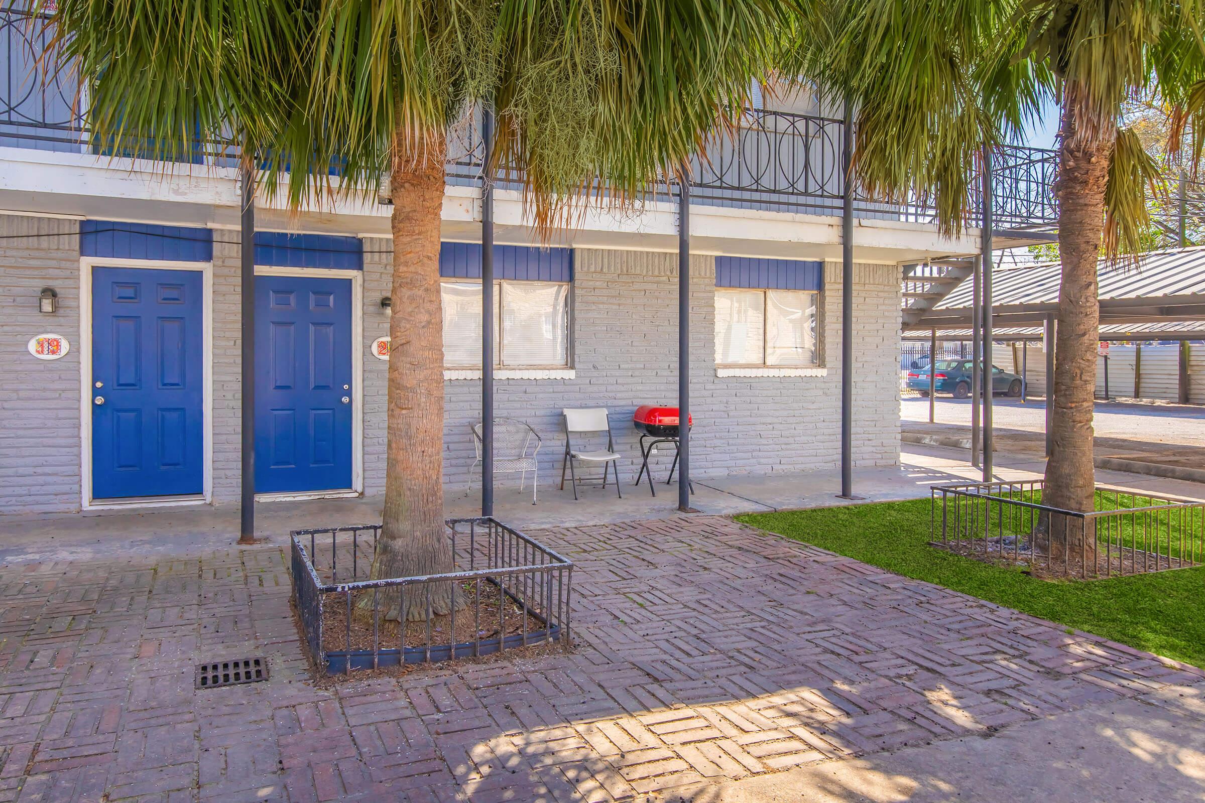 Two blue doors of a building are framed by palm trees, with a small patio area featuring two white chairs and a small red grill. The exterior is light gray brick, with windows featuring blue awnings. A paved walkway and green grass add to the setting, creating a cozy outdoor atmosphere.