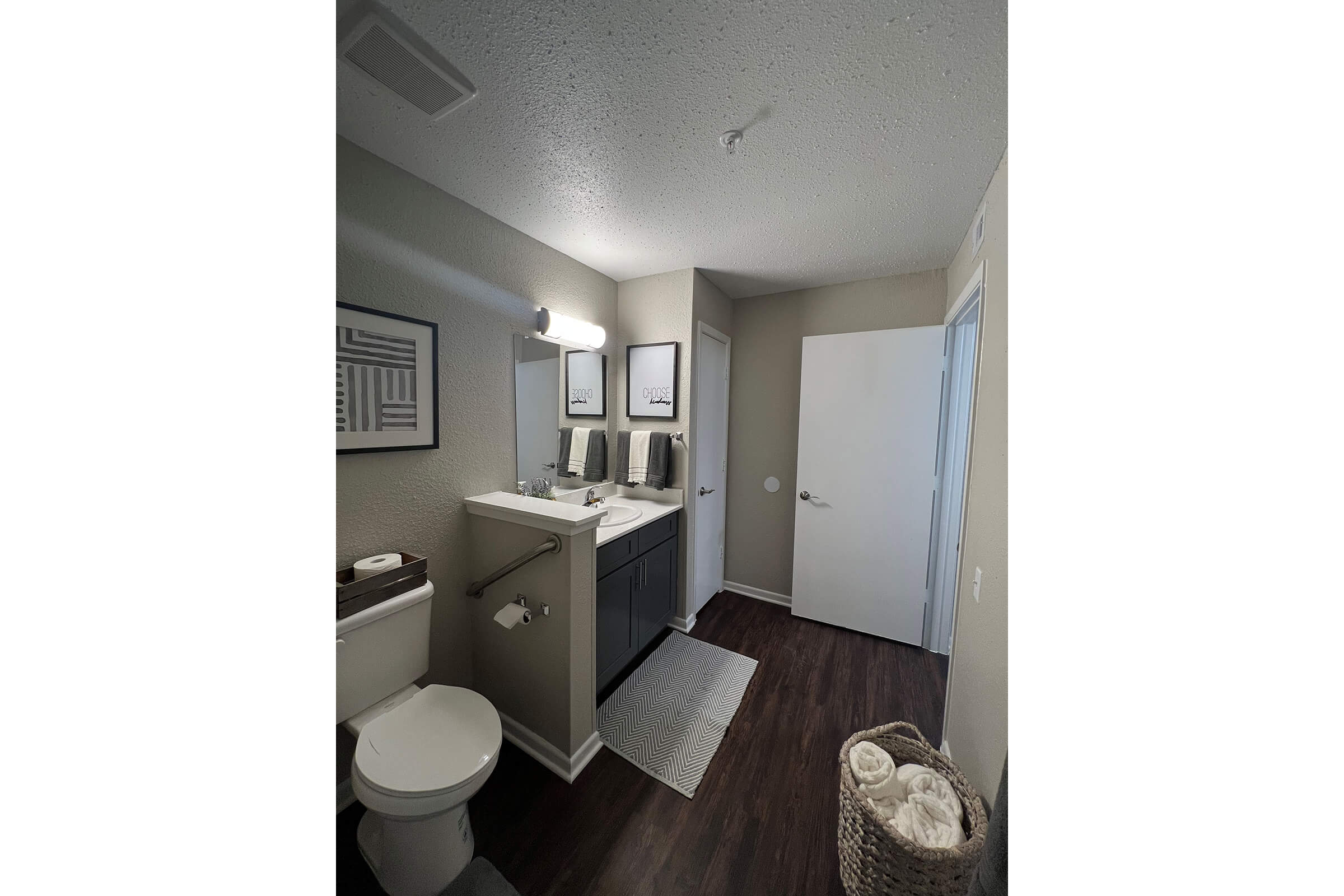 A modern bathroom featuring a vanity with two sinks, a large mirror, and light fixtures above. There’s a toilet, a textured rug on the floor, and a decorative basket with rolled towels. The walls are painted in a neutral tone, and the flooring is dark wood. A door leads to another space, and there are framed prints on the wall.