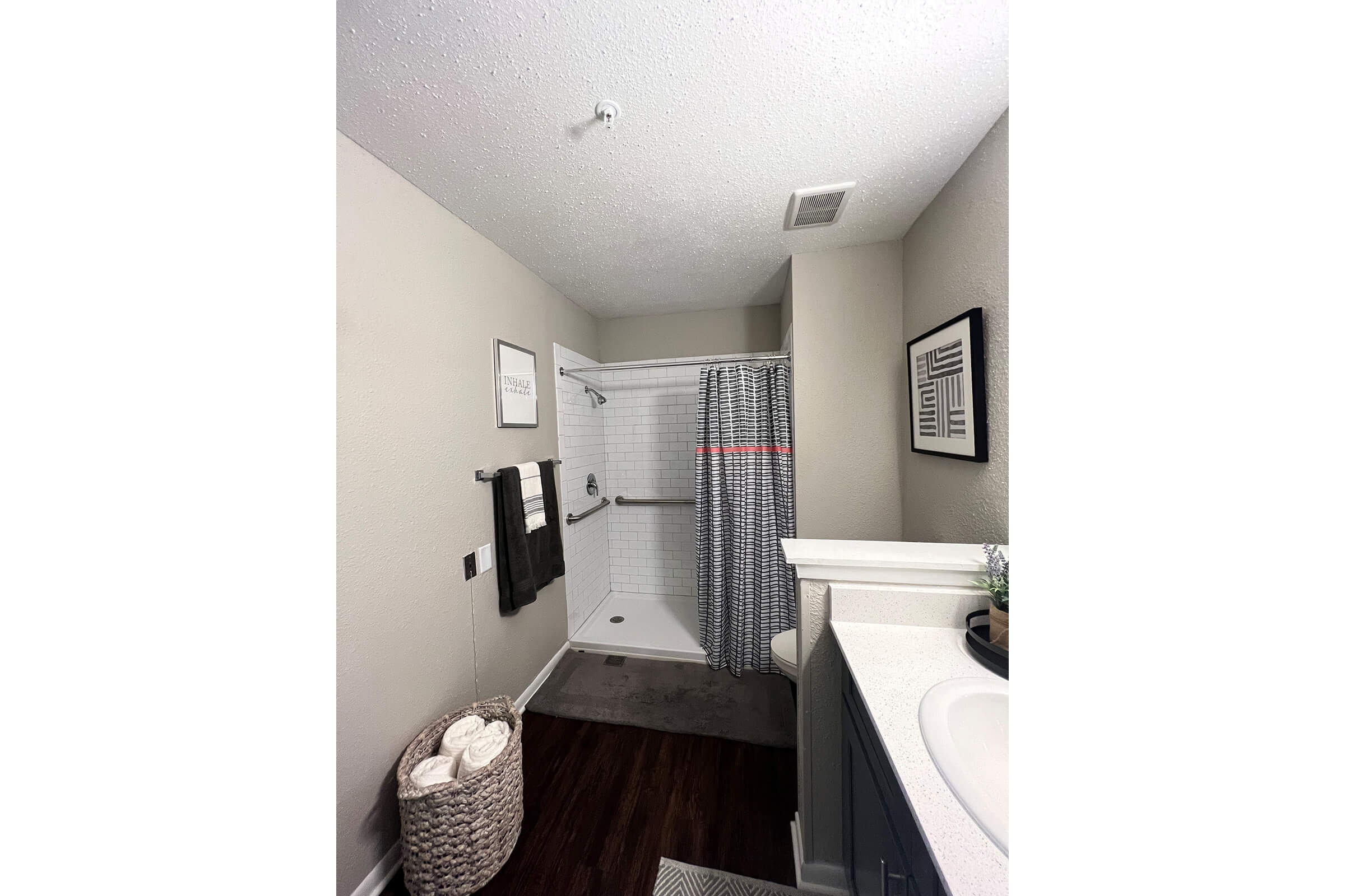 A clean and modern bathroom featuring a shower with a gray and white patterned curtain, a light-colored wall, and dark wood flooring. A countertop with a sink is on the right, accompanied by a storage basket filled with towels. Wall decor includes framed prints, enhancing the contemporary feel of the space.