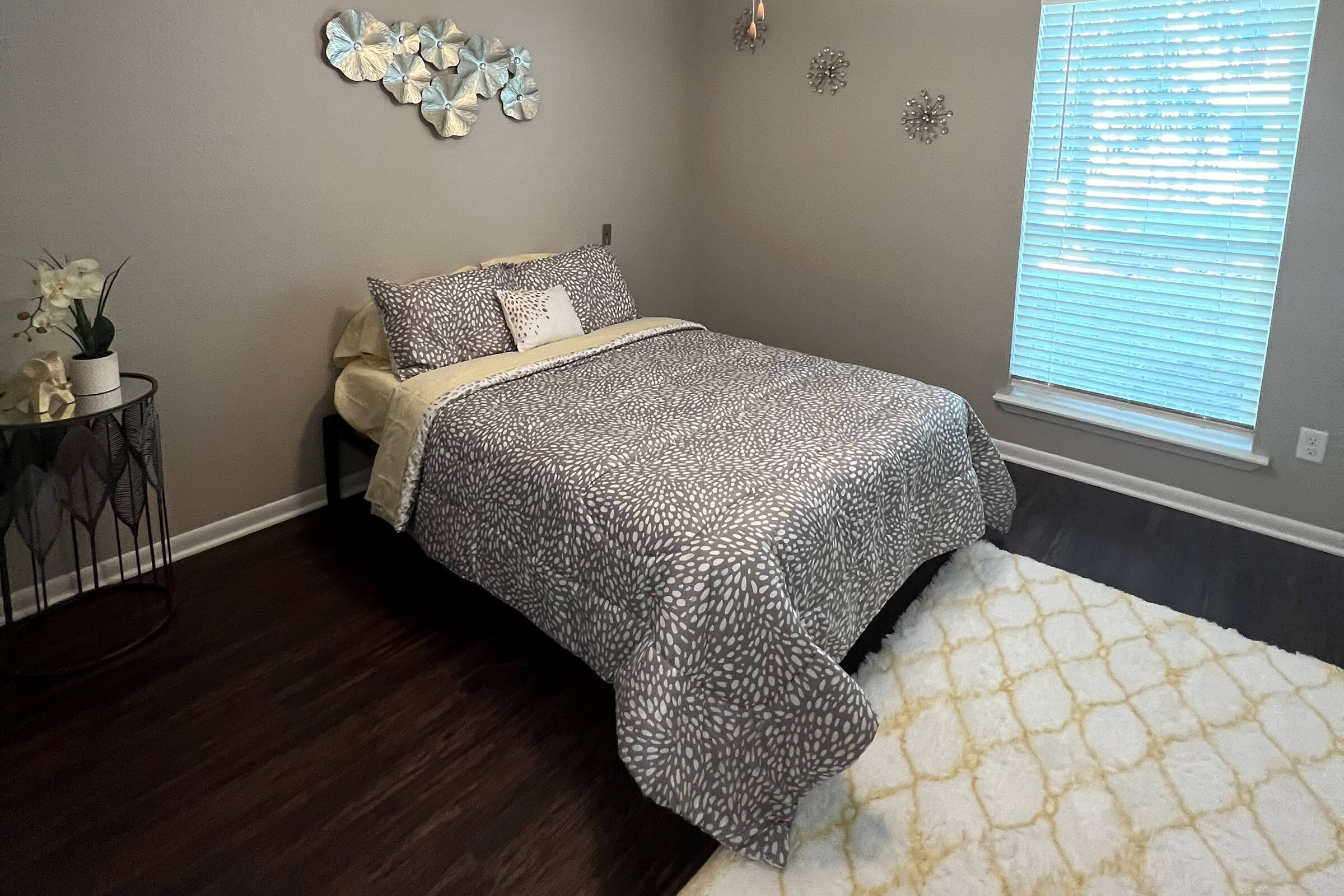 A cozy bedroom featuring a neatly made bed with a patterned gray comforter and decorative pillows. There are modern wall decorations above the bed, a small nightstand with a plant, and a large window covered with blinds that lets in soft natural light. A plush area rug adds warmth to the space.