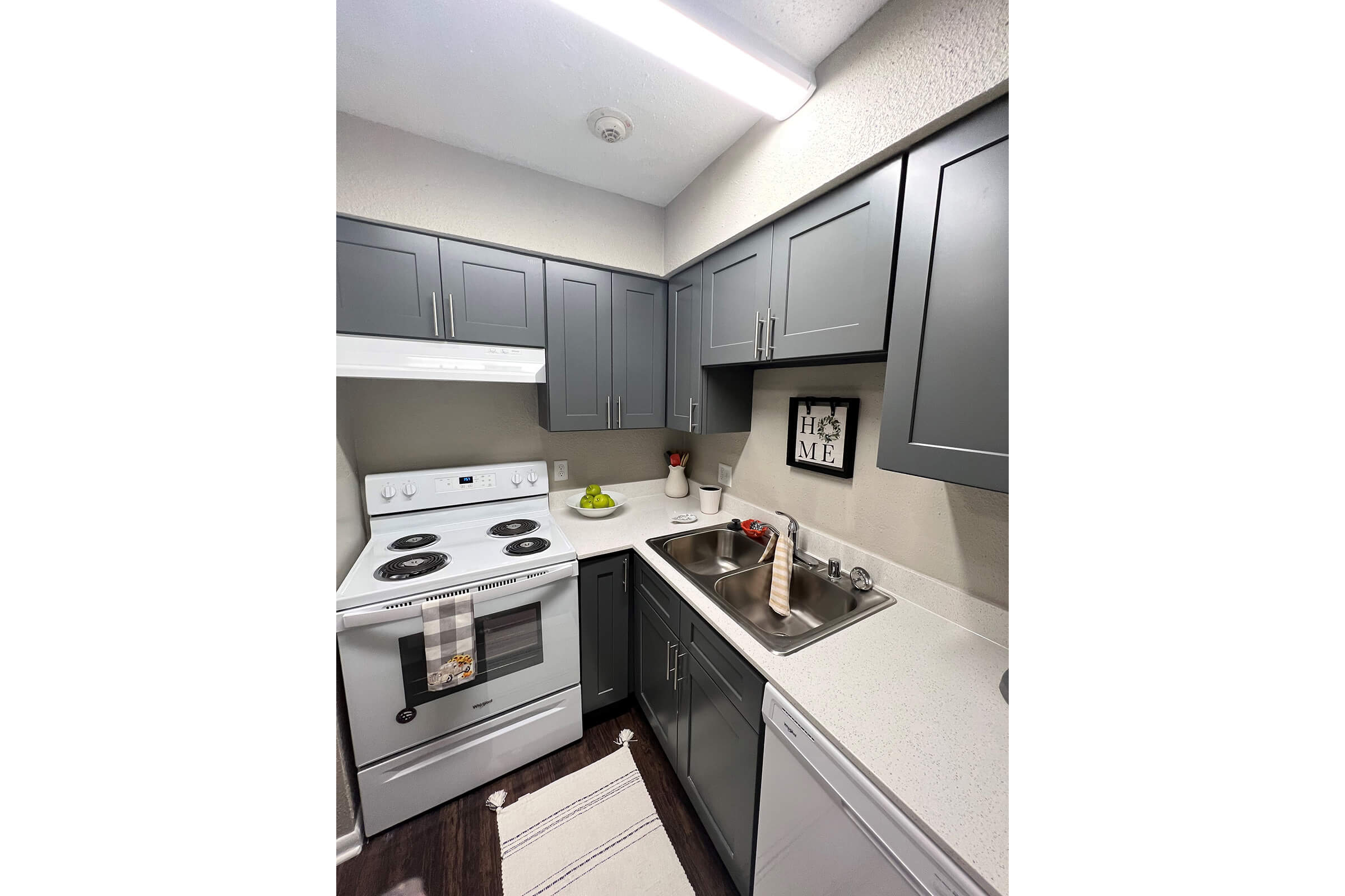 A small, modern kitchen featuring gray cabinets, a white stove, and a stainless-steel sink. The countertop is light-colored with a few decorations, including a bowl of green apples and a framed picture on the wall. Natural light illuminates the space, creating a cozy atmosphere.