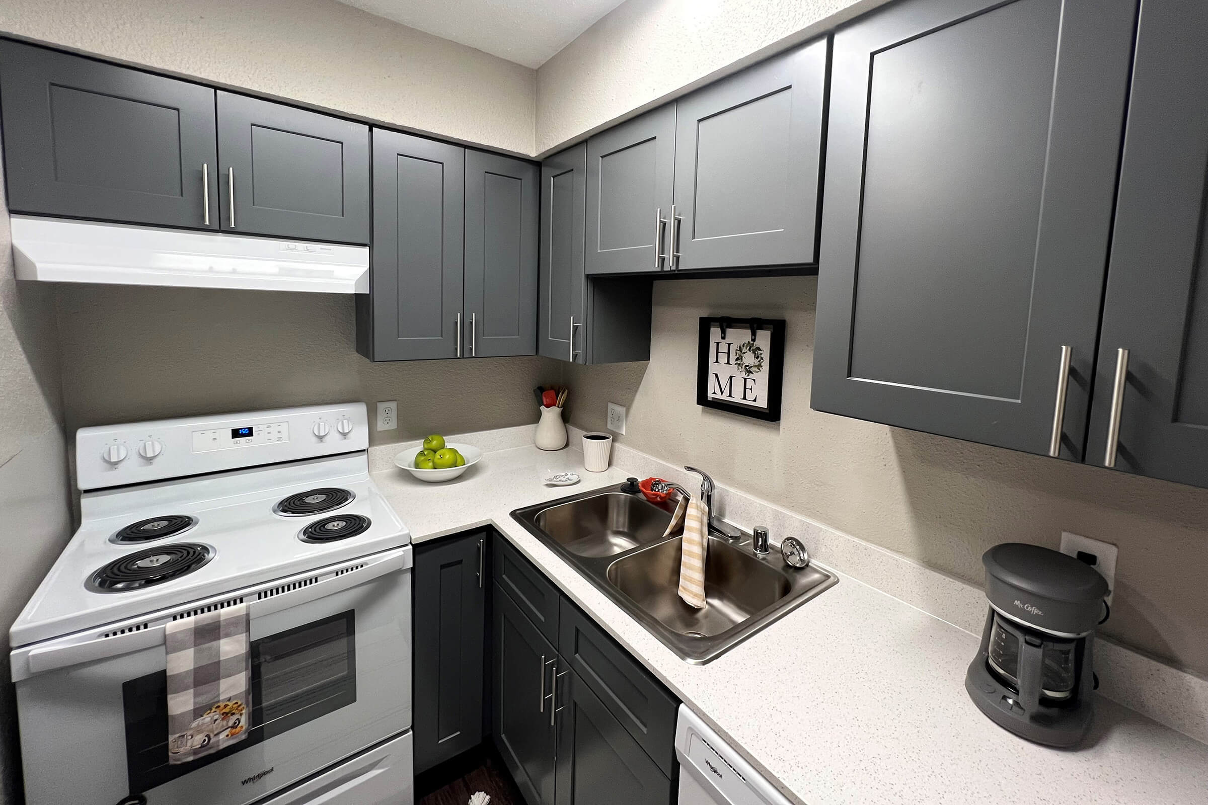 Modern kitchen featuring gray cabinets, white countertop, and stainless steel sink. The stove is white with four burners, and there’s a coffee maker on the counter. A decorative sign that reads "HOME" is displayed on the wall, alongside a small bowl of green apples and a vase with flowers.