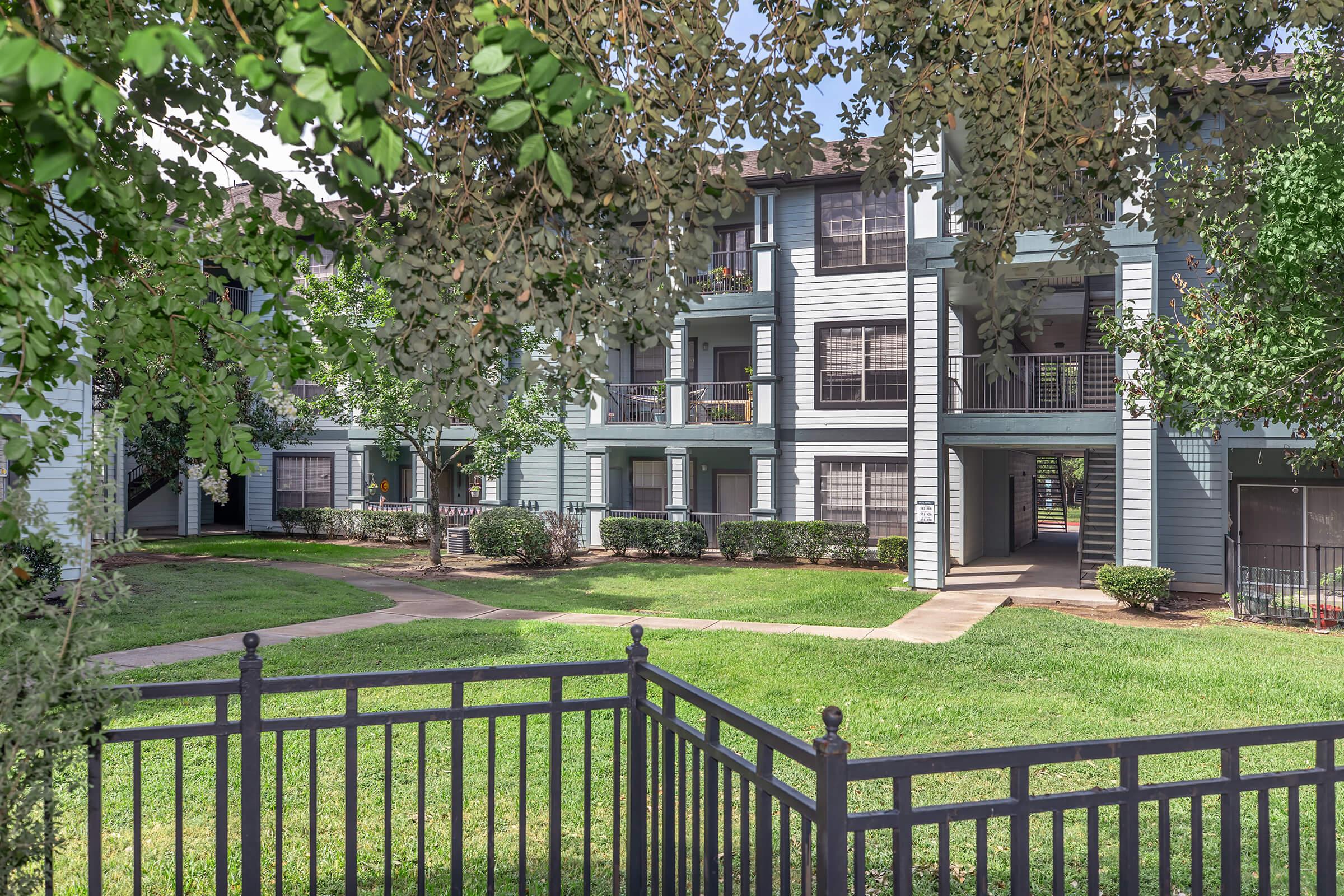 A view of a multi-unit residential apartment complex surrounded by green grass and trees. The building features blue siding and several balconies. A pathway leads through the grass, with a decorative black wrought-iron fence in the foreground. The setting is bright and inviting, suggesting a well-maintained living environment.