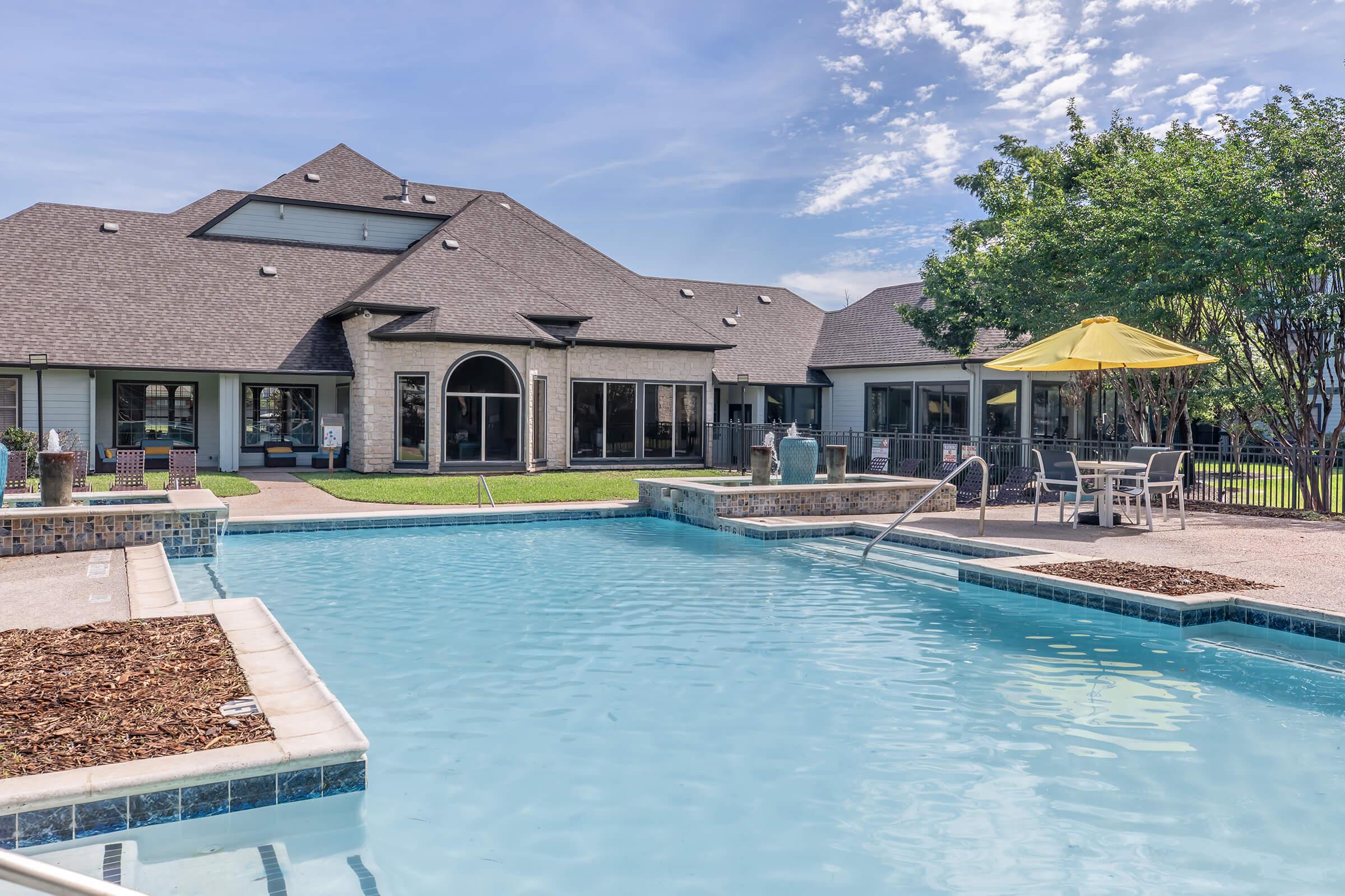 A bright outdoor pool area featuring a shimmering blue pool with a small spa, surrounded by well-maintained landscaping. In the background, a spacious building with large windows and a patio area includes outdoor furniture and an umbrella for shade. The sky is clear with a few clouds.