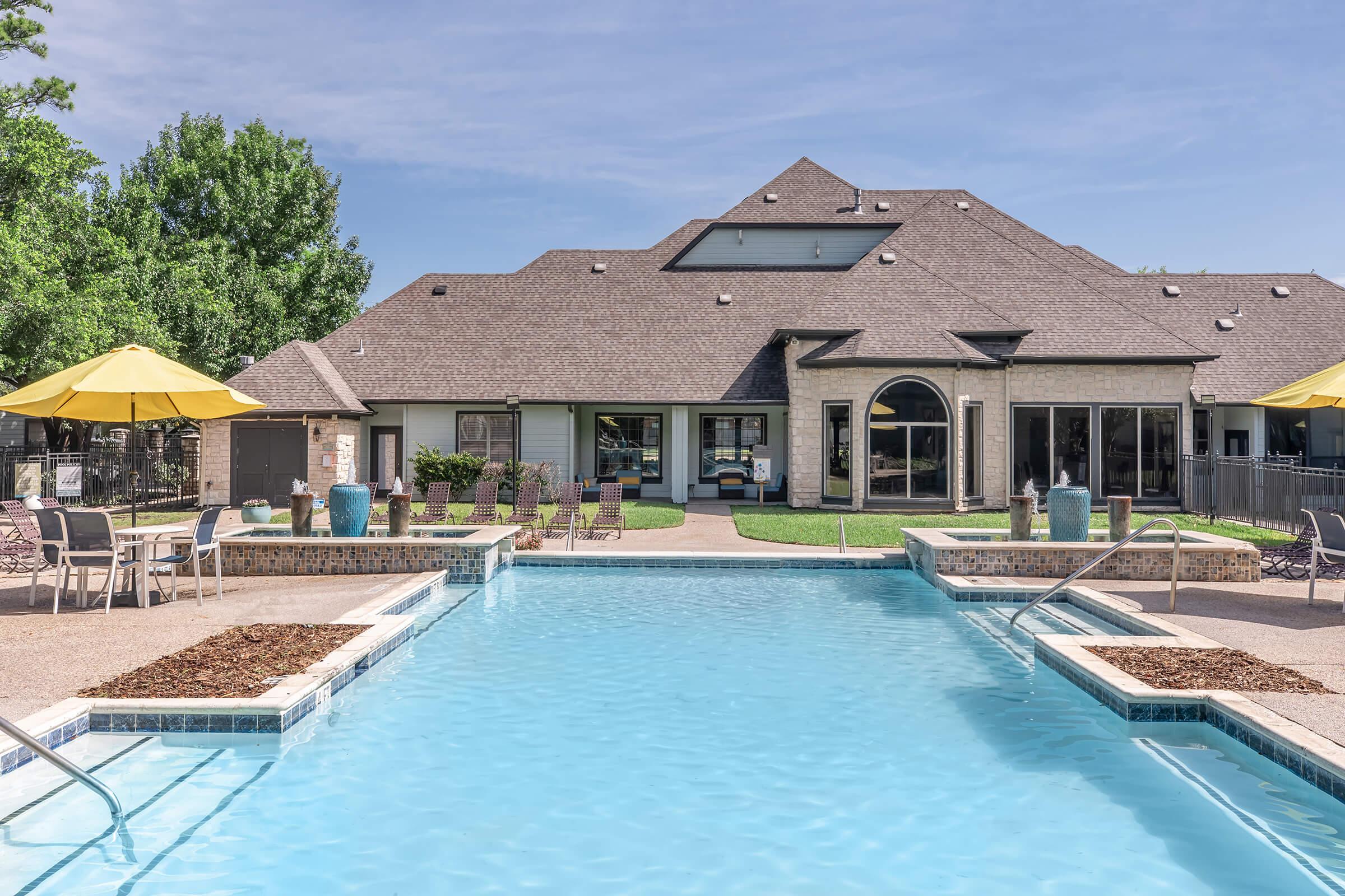A clear blue swimming pool with steps leading in, surrounded by lounge chairs and umbrellas. In the background, a large, modern building features arched windows and a well-maintained lawn. The scene is bright and inviting, suggesting a relaxing outdoor space.