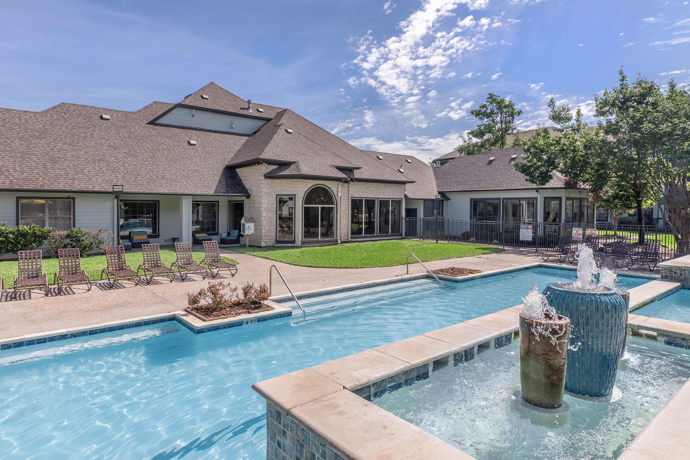 A serene outdoor area featuring a swimming pool with a fountain, surrounded by lounge chairs and a well-maintained lawn. In the background, there are modern buildings with large windows, under a bright blue sky with a few clouds. Ideal for relaxation and leisure activities.