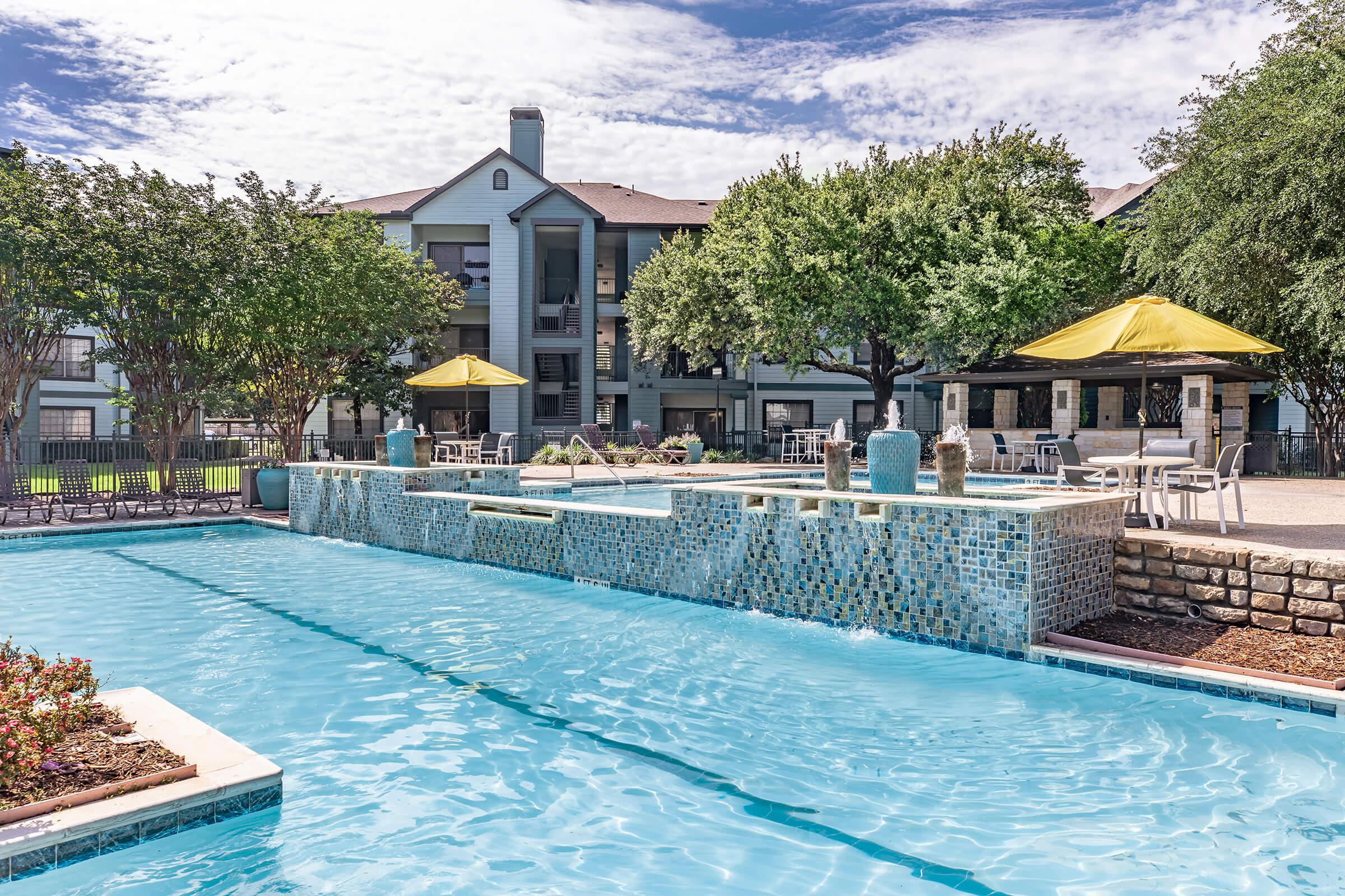 A serene outdoor pool area featuring clear blue water, bordered by lush green trees and landscaped flower beds. In the background, a modern apartment building can be seen, with yellow umbrellas providing shade over tables nearby. The setting is bright and inviting, perfect for relaxation.
