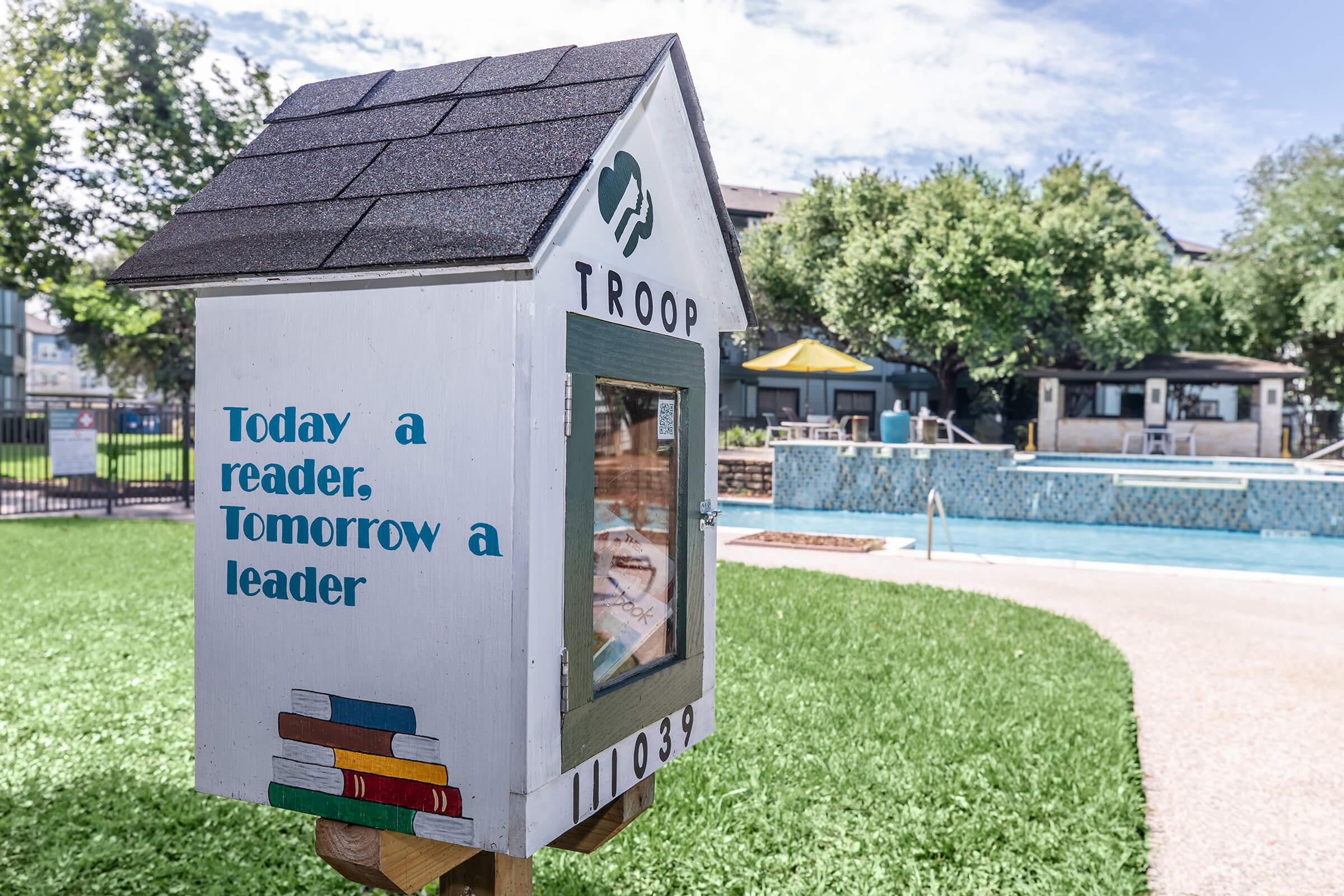 A small, colorful wooden library box titled "TROOP" stands on a grassy area near a pool. The box features the text "Today a reader, Tomorrow a leader" and a graphic of stacked books. In the background, there are trees and a pool area, indicating a community space for reading and relaxation.