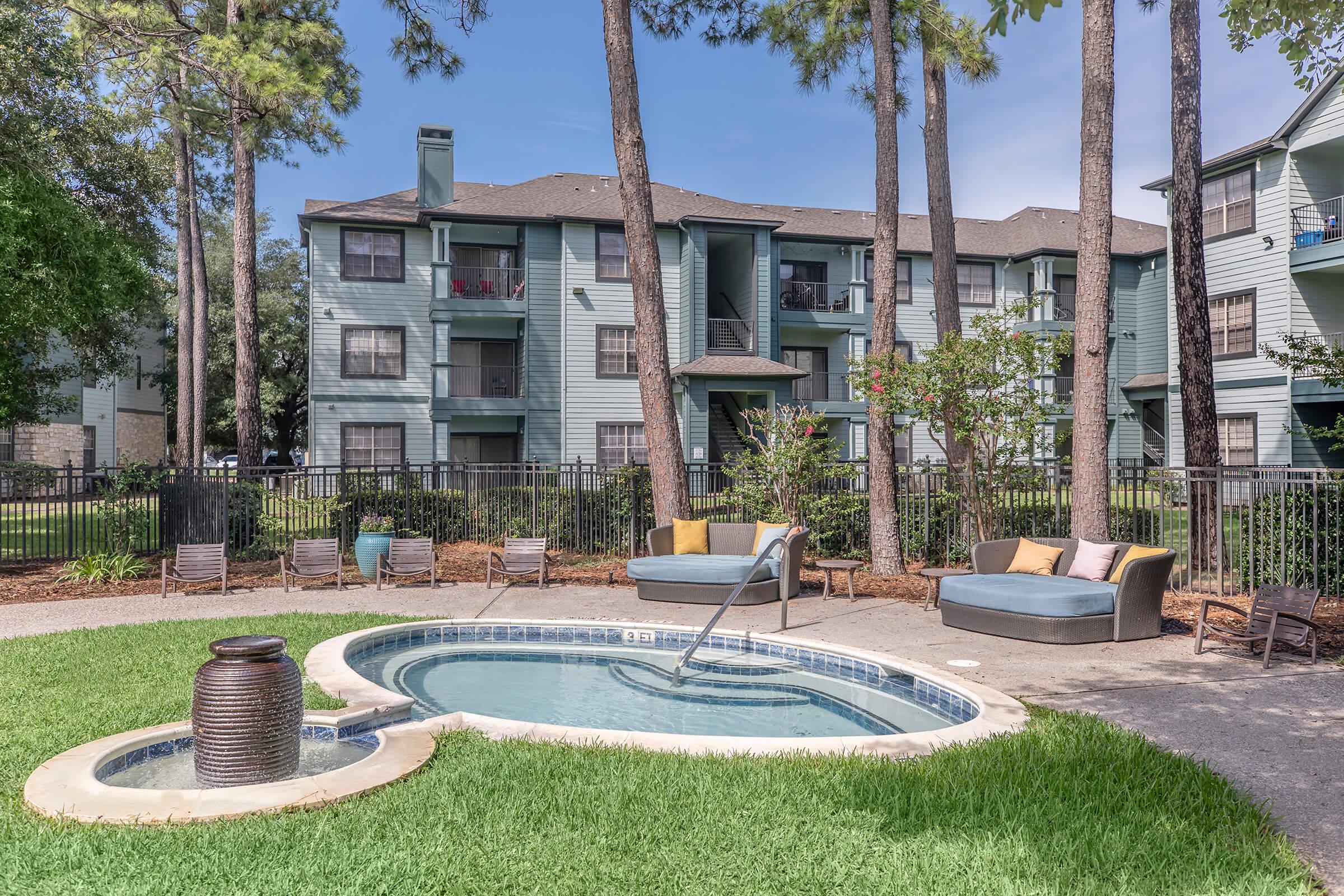 A serene outdoor area featuring a small spa or hot tub surrounded by a green lawn. In the background, a three-story apartment building painted in light blue, with several balconies visible. Comfortable seating arrangements and potted plants are placed around the pool area, under tall trees against a clear sky.