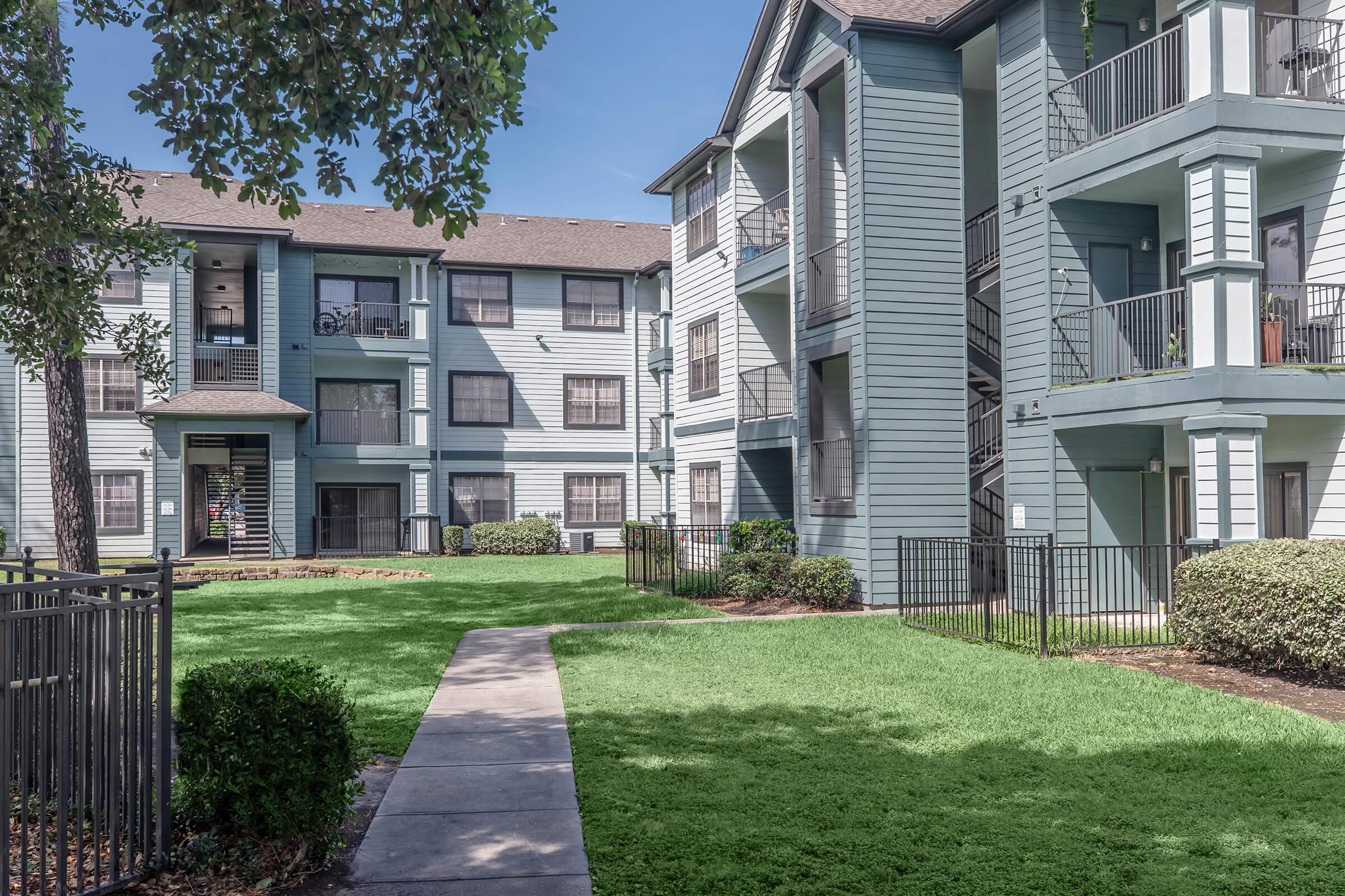 A view of a multi-story apartment complex with light blue exteriors, featuring balconies and black railings. In the foreground, a well-maintained green lawn and neatly trimmed bushes surround a paved path leading through the courtyard, providing a welcoming residential atmosphere.
