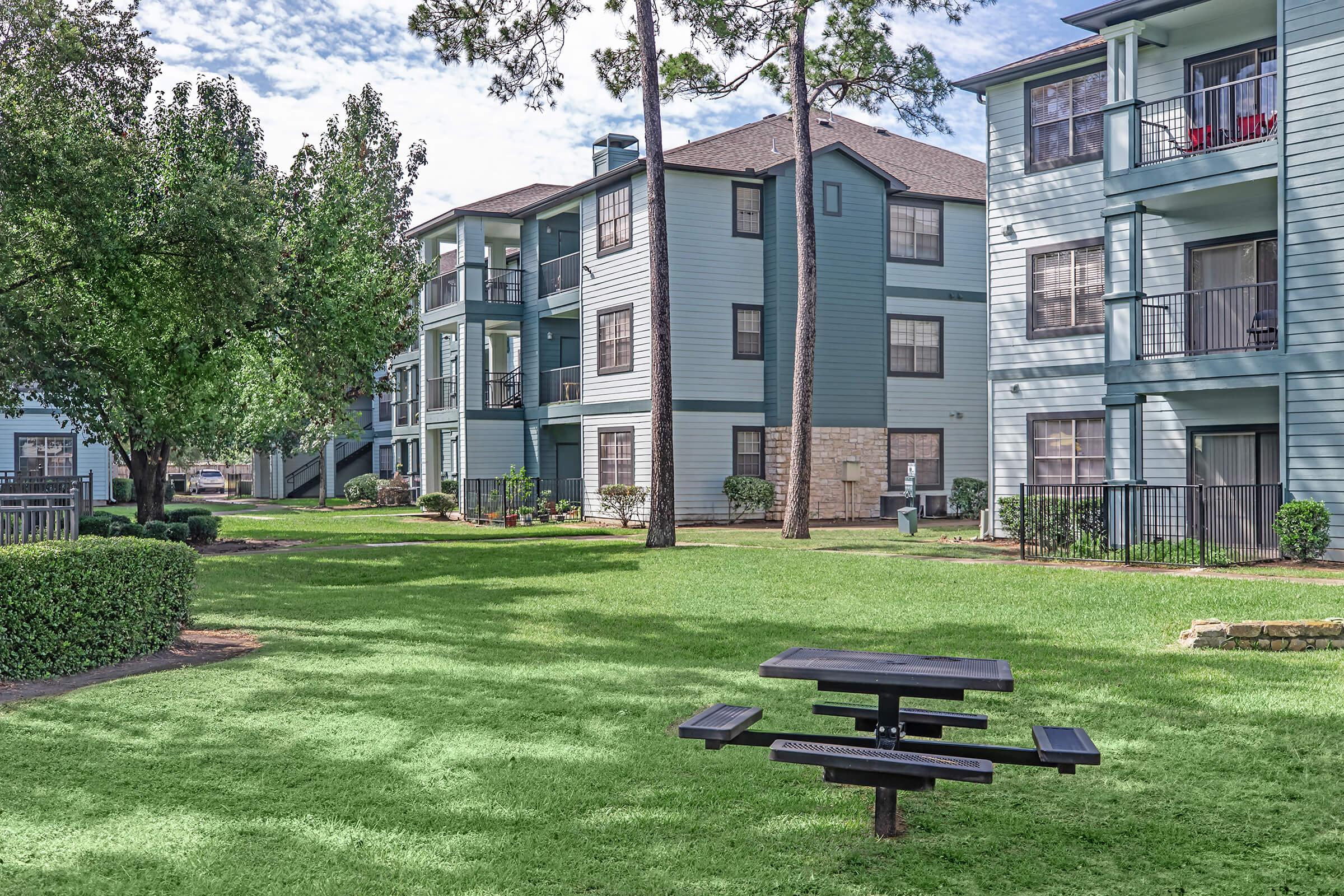 A residential apartment complex featuring multiple light blue buildings arranged around a green lawn. In the foreground, there is a black picnic table on the grass, surrounded by trees and shrubs. The scene captures a peaceful outdoor area with a clear sky overhead.