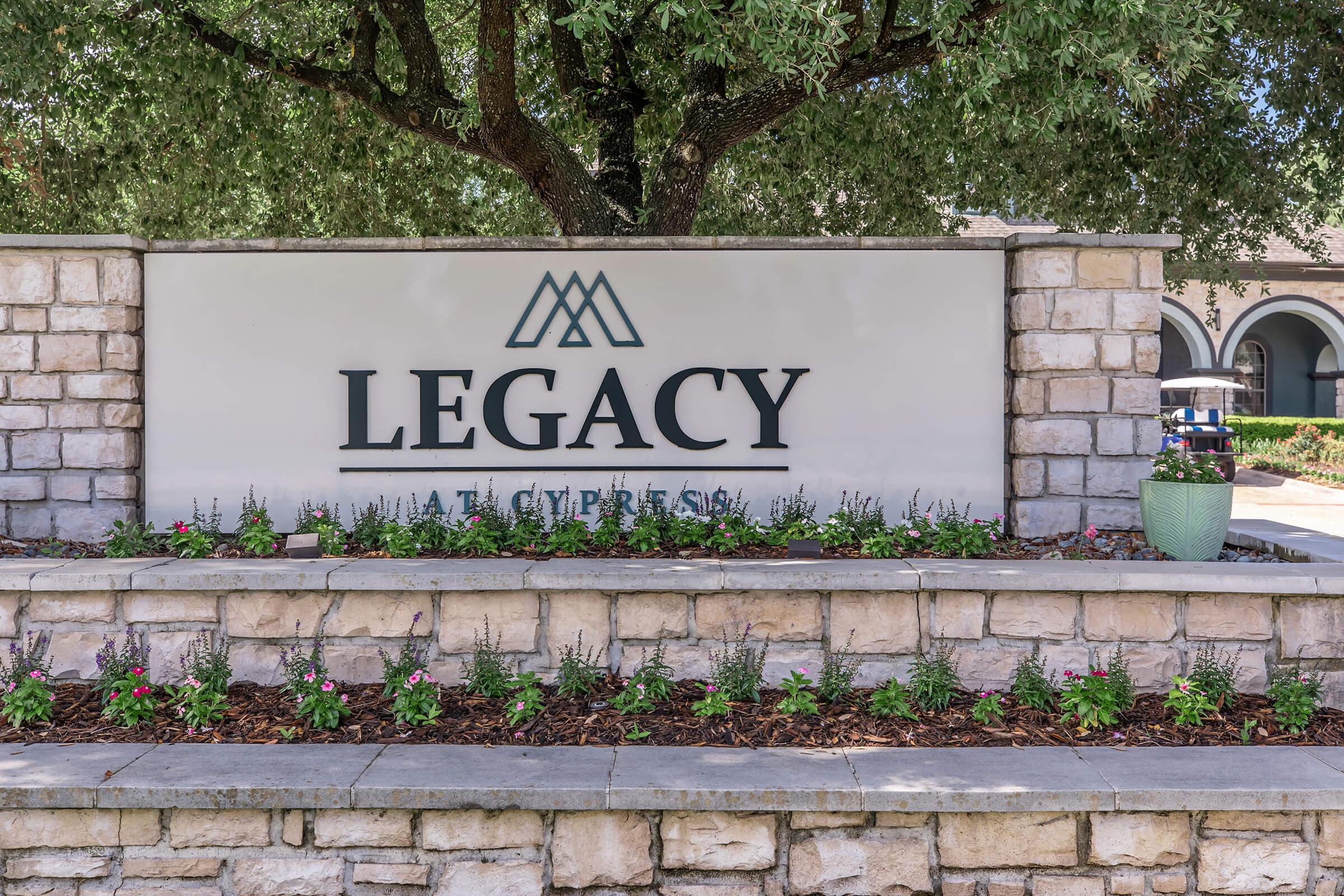 A stone wall featuring a large sign that reads "LEGACY AT CYPRESS" in bold black letters. The sign is surrounded by a decorative planter with flowers and is positioned under a large tree, providing a welcoming atmosphere.