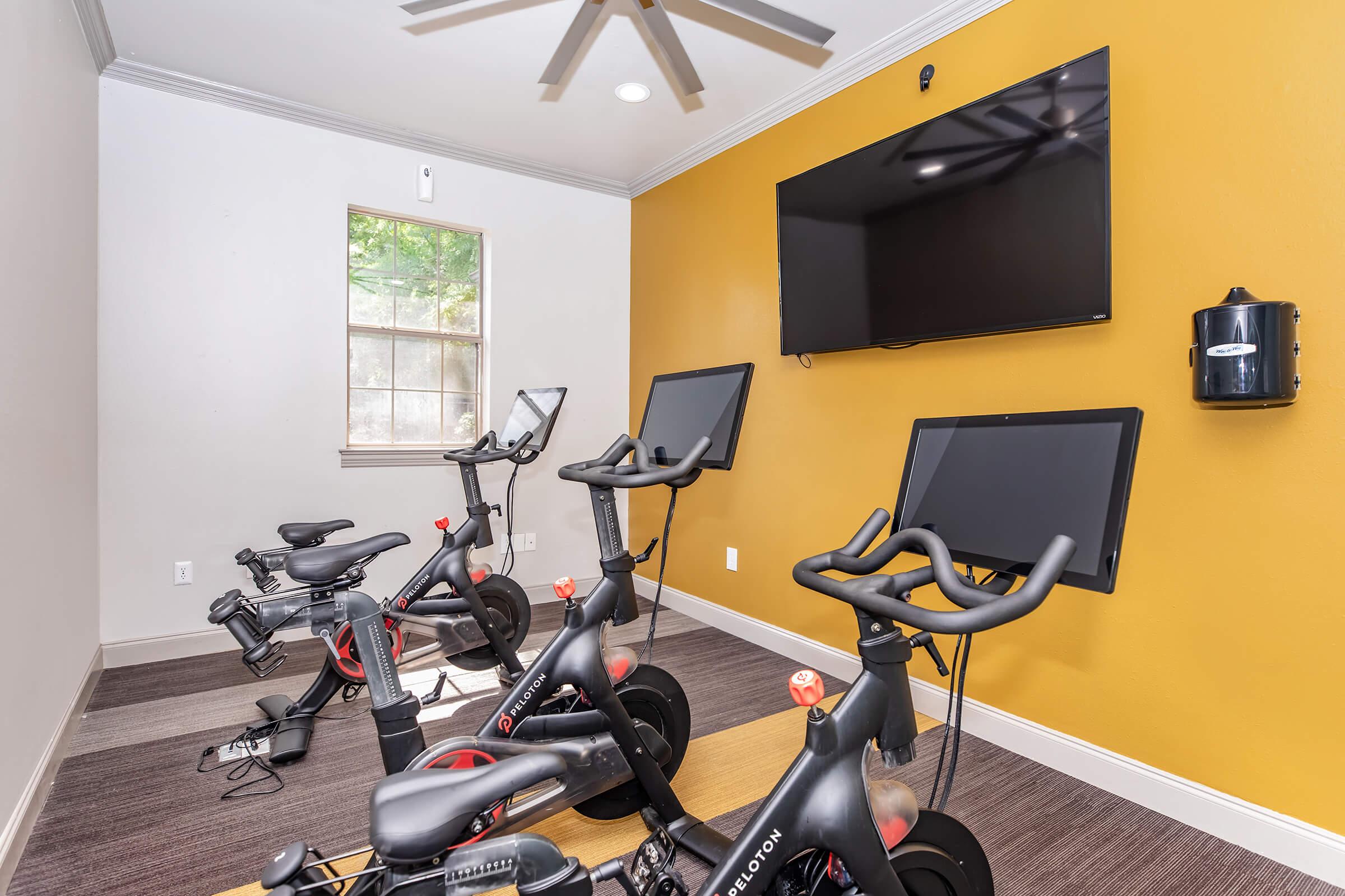 Indoor cycling studio featuring multiple stationary bikes with screens, a wall-mounted television, and a bright yellow accent wall. Natural light comes through a window, enhancing the workout space with a modern and inviting atmosphere.