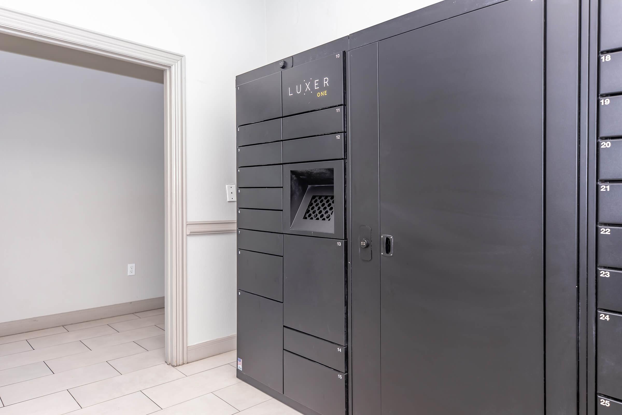 A black automated locker system labeled "LUXER" set in a well-lit room with beige tile flooring. An open doorway is visible in the background leading to another area. The lockers have multiple compartments numbered from 1 to 26, with a mesh ventilation area on one section.