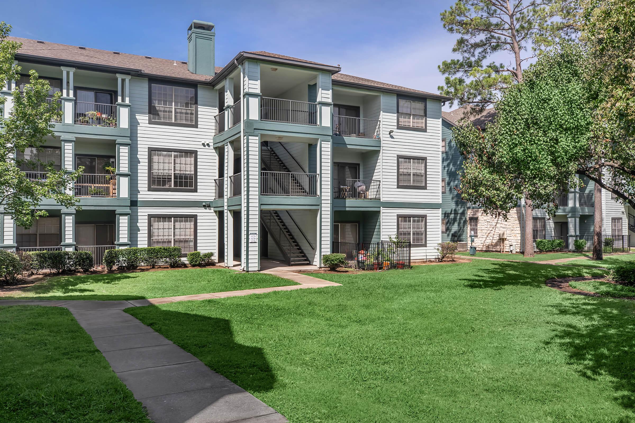 A well-maintained apartment complex featuring three stories, with a light blue exterior. The building has balconies, decorative plants, and a grassy area in front. Pathways lead through the landscaped grounds, providing a pleasant outdoor space. Bright, sunny skies complement the scene.