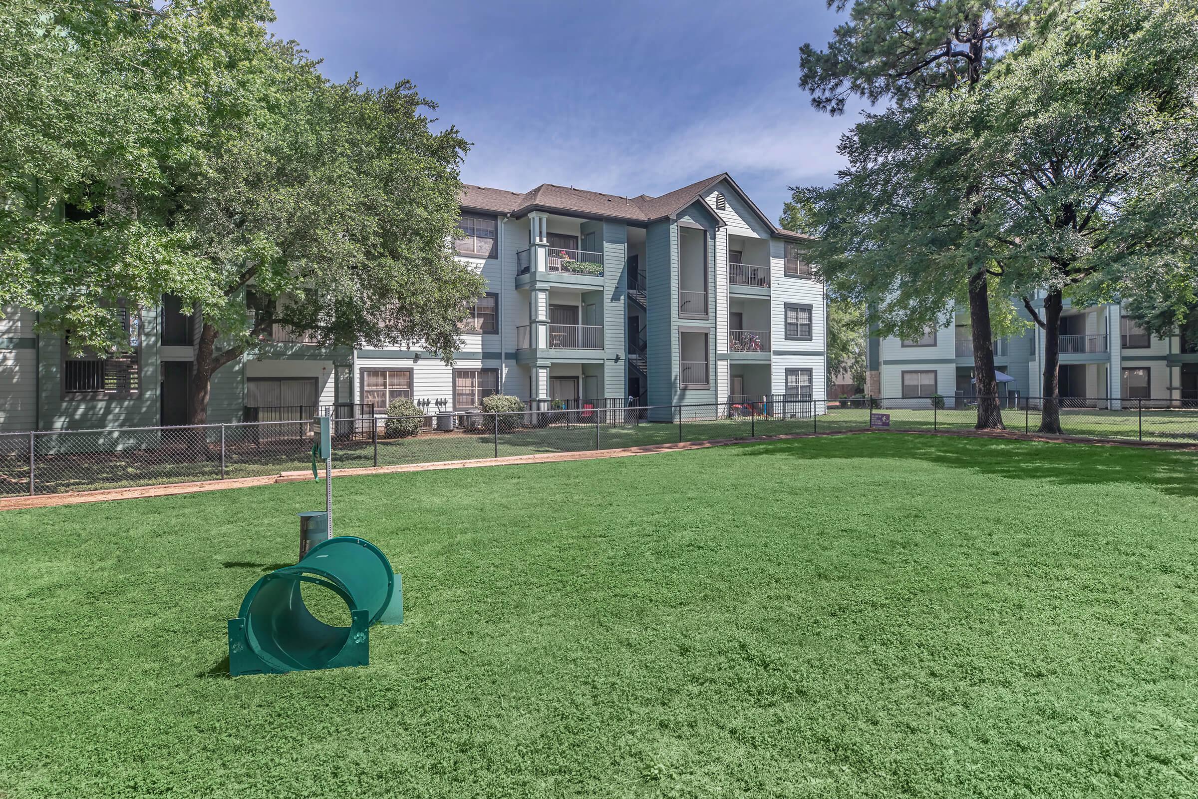 View of a green lawn area featuring a blue slide, surrounded by a fence and residential apartment buildings. Trees provide shade, and the sky is clear with a hint of clouds. The setting appears to be well-maintained and inviting for outdoor activities.