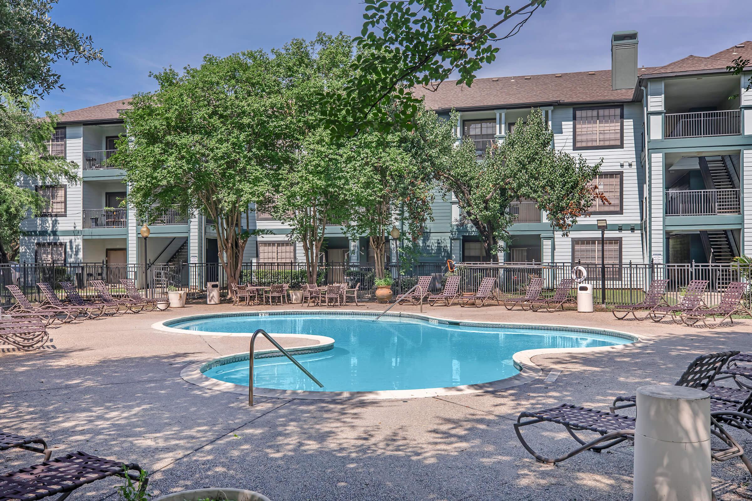 A tranquil outdoor pool area surrounded by lounge chairs, with lush green trees and a multi-story residential building in the background. The pool is clear and inviting, set in a serene atmosphere perfect for relaxation.